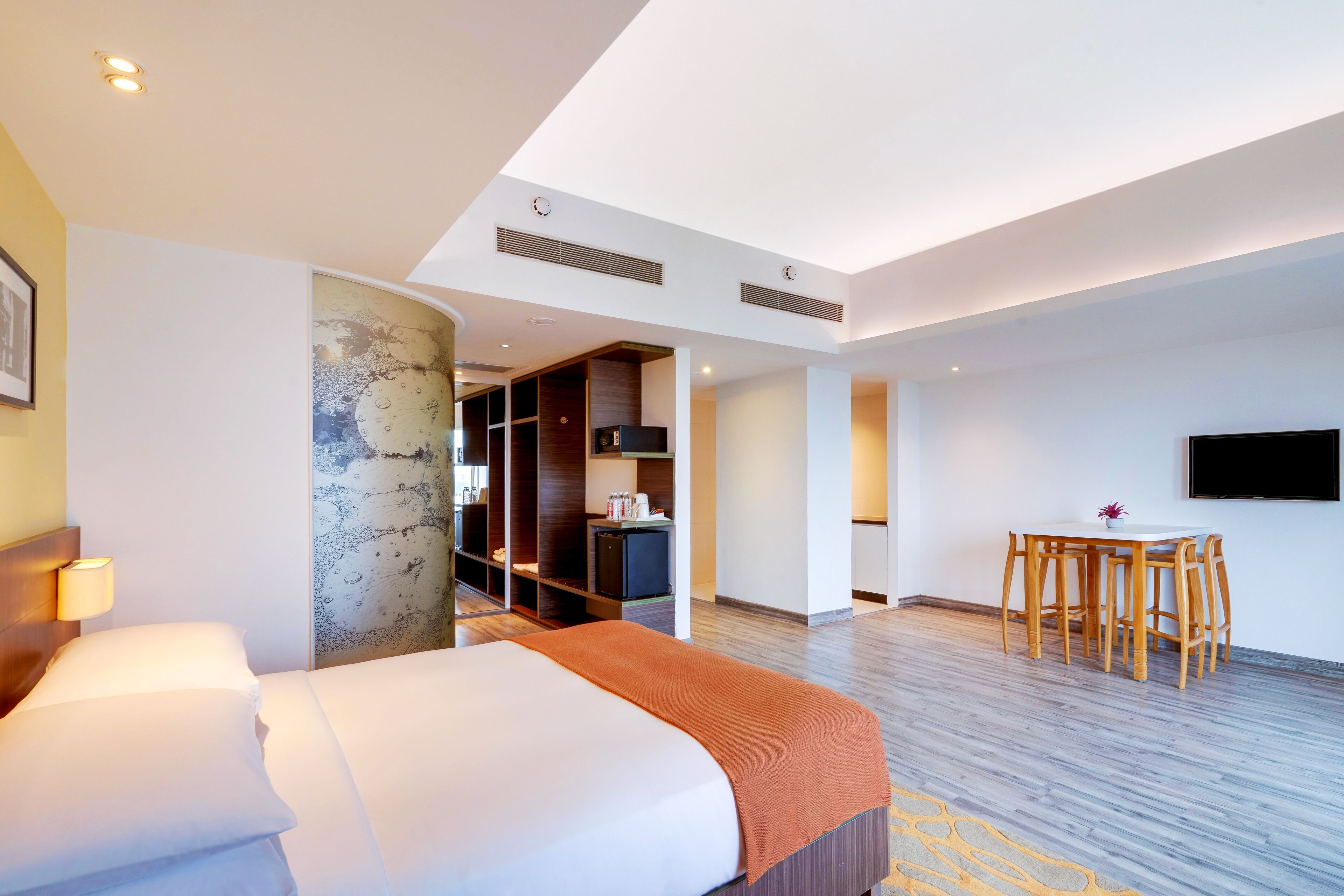 A hotel room with a bed, a dining table with chairs, a TV mounted on the wall, and a kitchenette area.