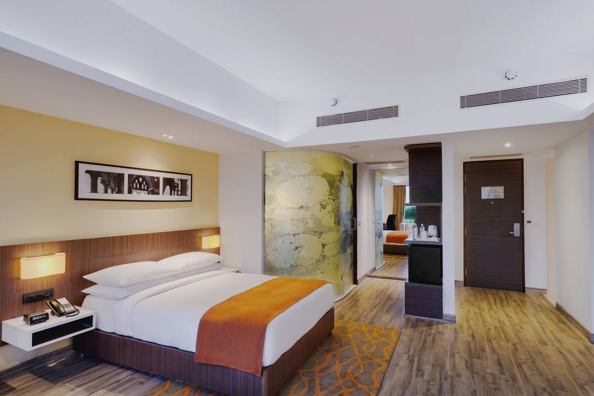 Ein Hotelzimmer mit Bett, Nachttisch, Lampe, Kunstwerk und einem durch eine Glastrennwand einsehbaren Badezimmer.