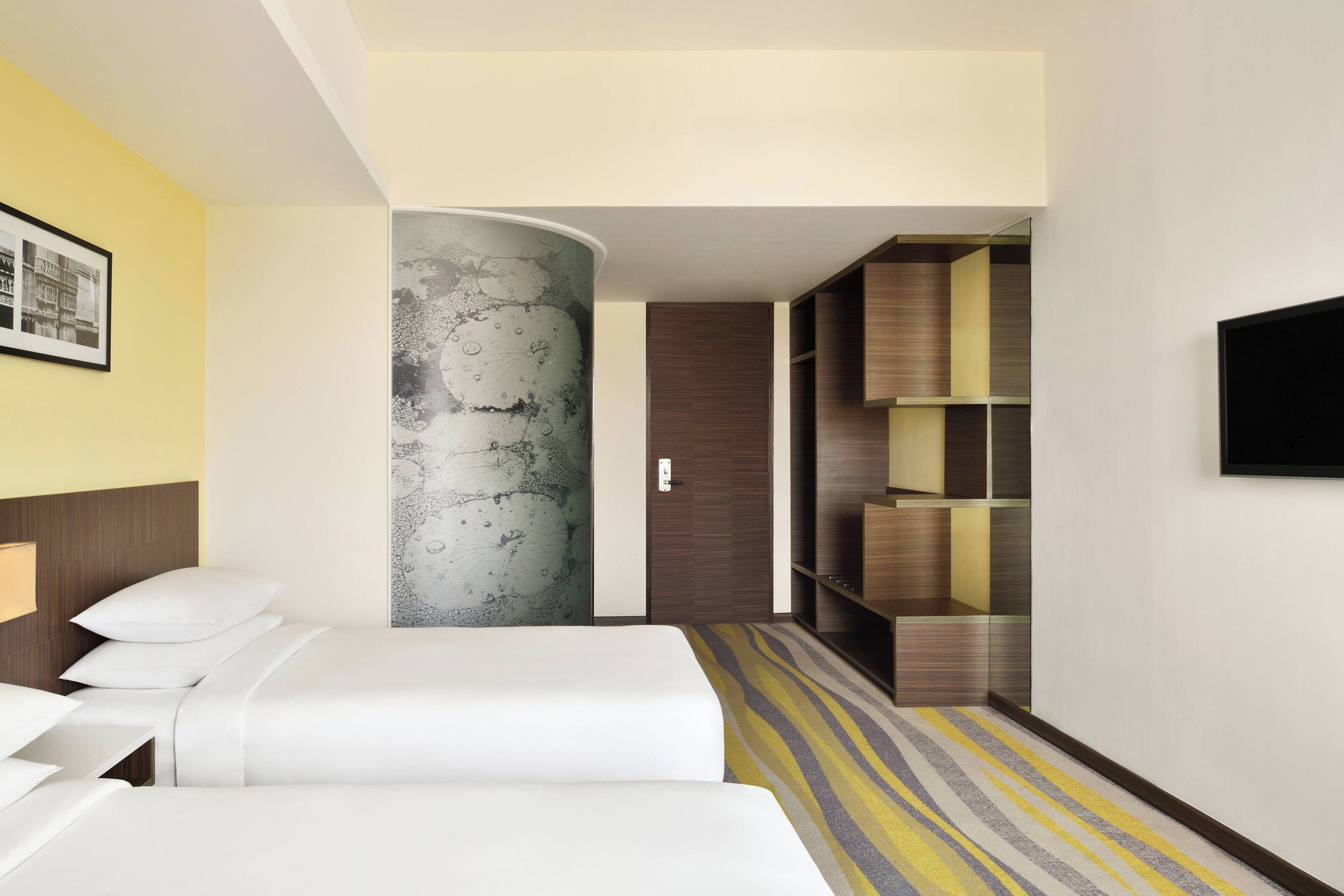 A hotel room with two beds, a large abstract wall art, a wardrobe, and a television on the wall.