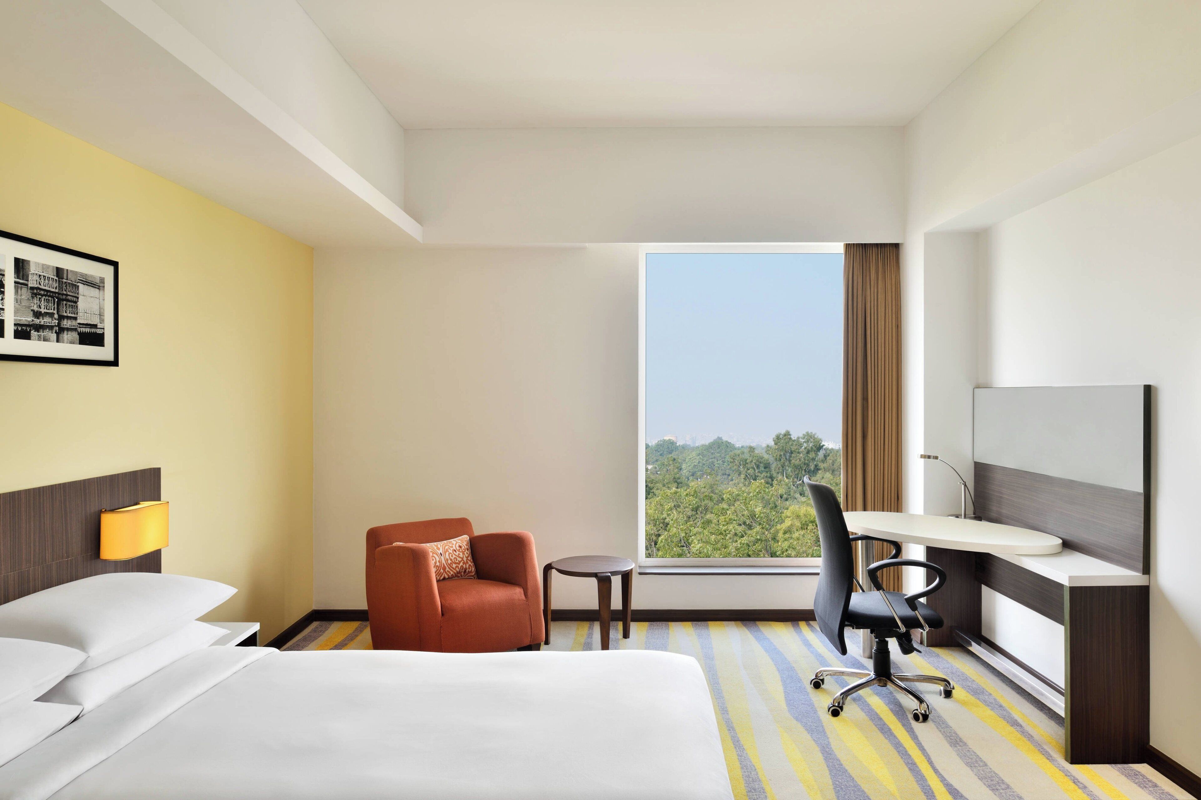 A hotel room with a bed, a desk, a chair, a small table, and a window with a view of greenery.