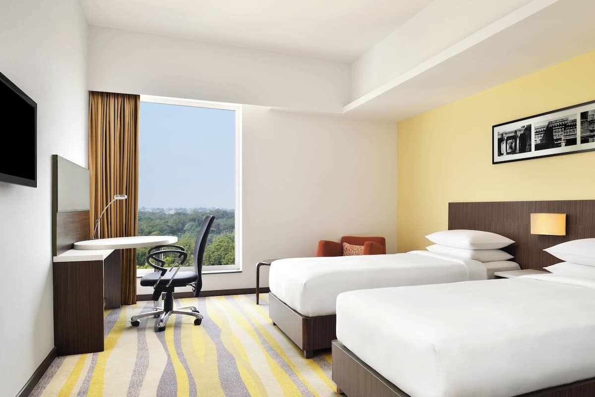 Ein Hotelzimmer mit zwei Betten, einem Schreibtisch, einem Sessel, einem Fenster mit Ausblick und einem an der Wand befestigten Fernseher.
