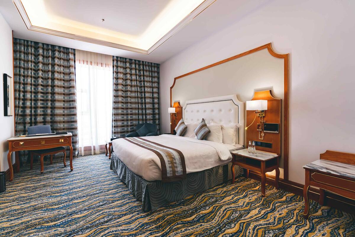 A hotel room with a large bed, a desk, and a chair. There is a window with curtains and a wall-mounted TV.