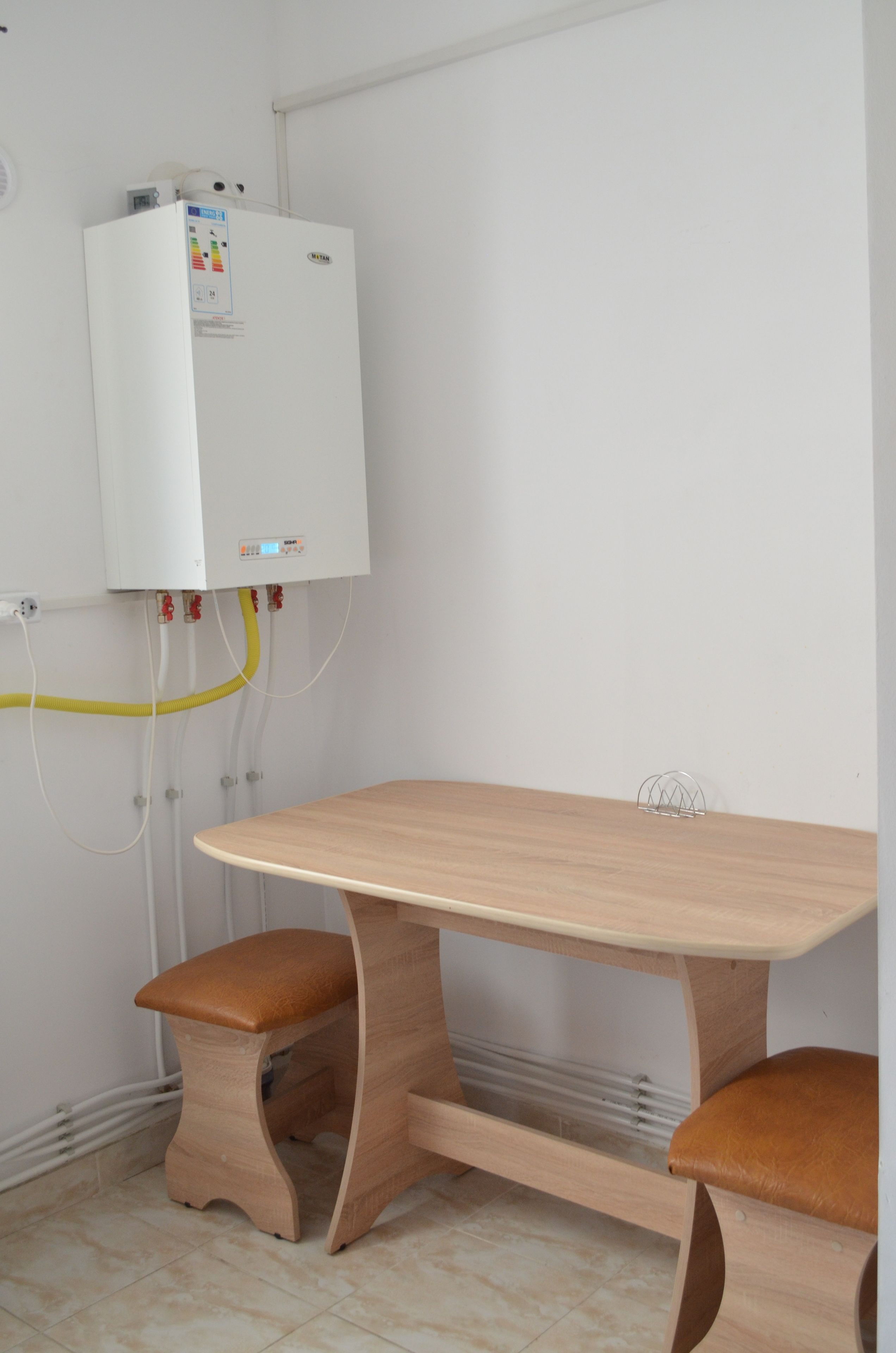 A room with a wooden table and two stools, a white wall-mounted heater, and visible plumbing.