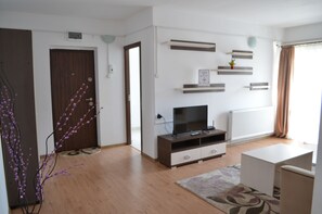 A modern living room with a wooden floor, a TV on a wooden stand, a white sofa, and a small table. There are decorative shelves on the wall and a door with a wooden frame. - Alexys Residence 6 (Iasi)