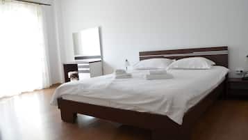 Cama bem arrumada com lençóis e toalhas brancas, cabeceira de madeira, mesa de cabeceira com abajur e um espelho grande na parede.