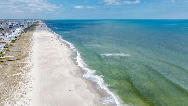 Une vue aérienne d’une plage avec une longue étendue de sable, quelques personnes et des bâtiments le long du rivage.
