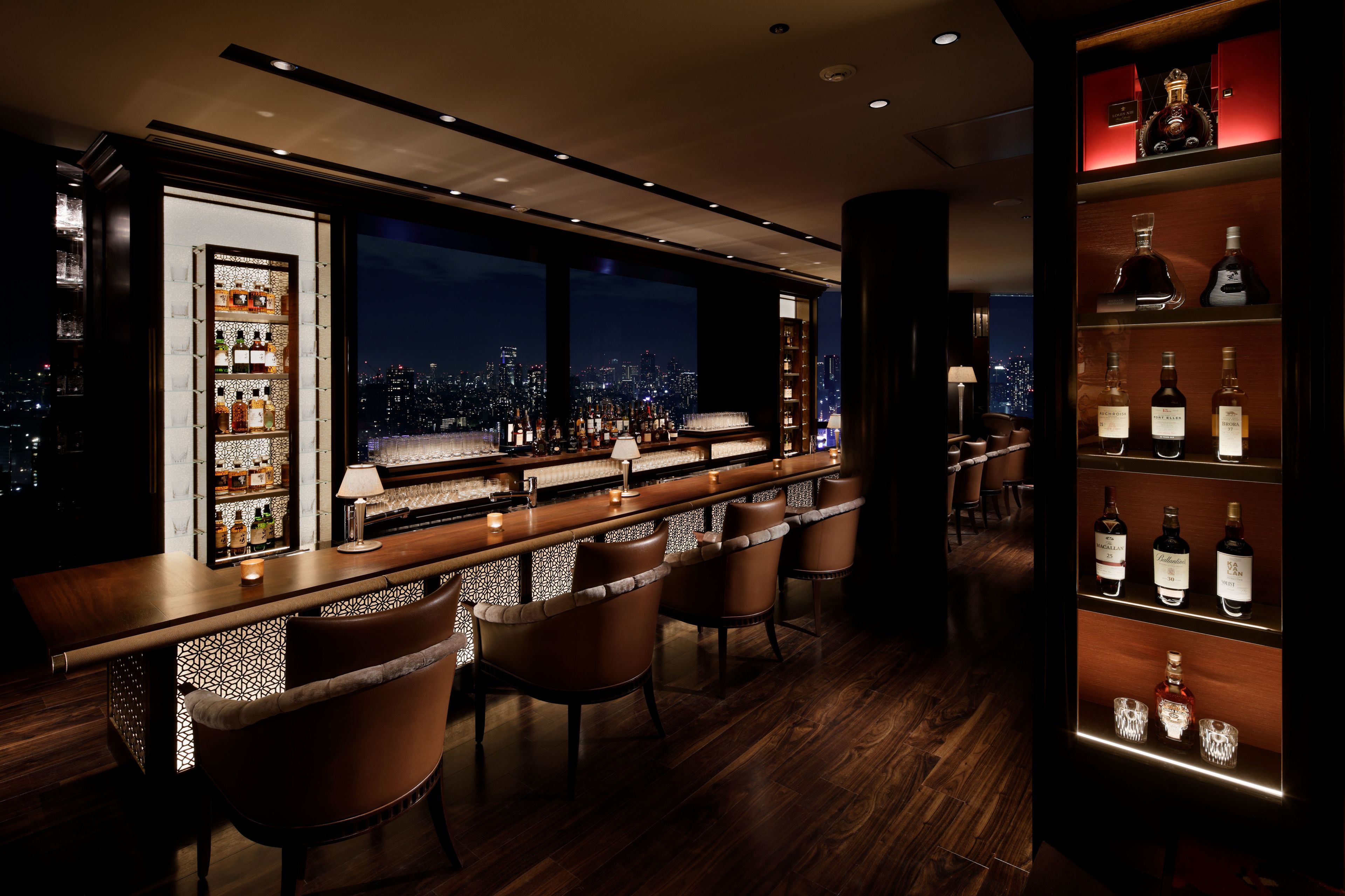 Zona de bar con vistas a la ciudad, barra de madera, sillas de cuero y estantes con botellas.