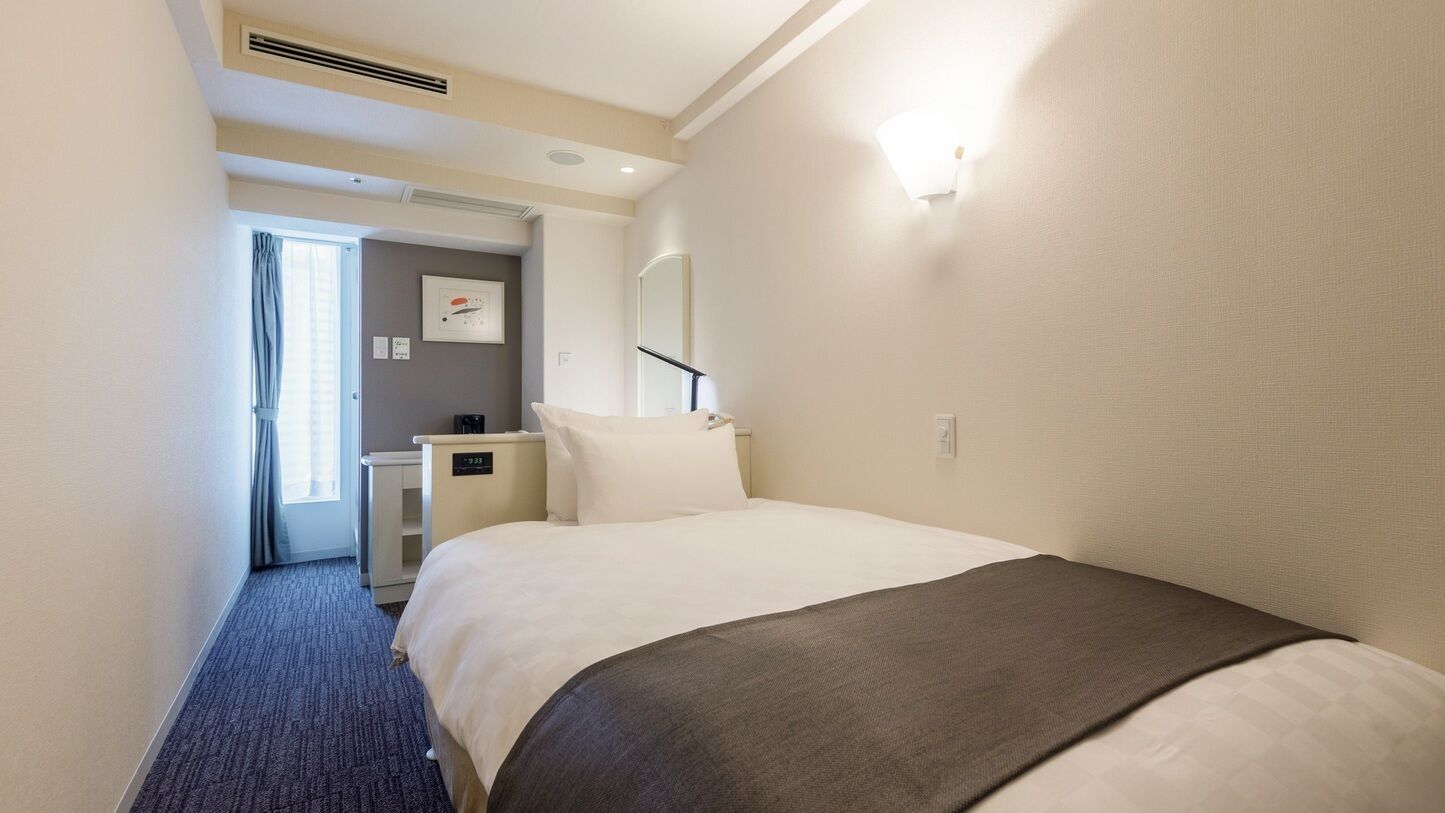 A hotel room with a bed, a desk, a chair, a mirror, and a window with curtains.