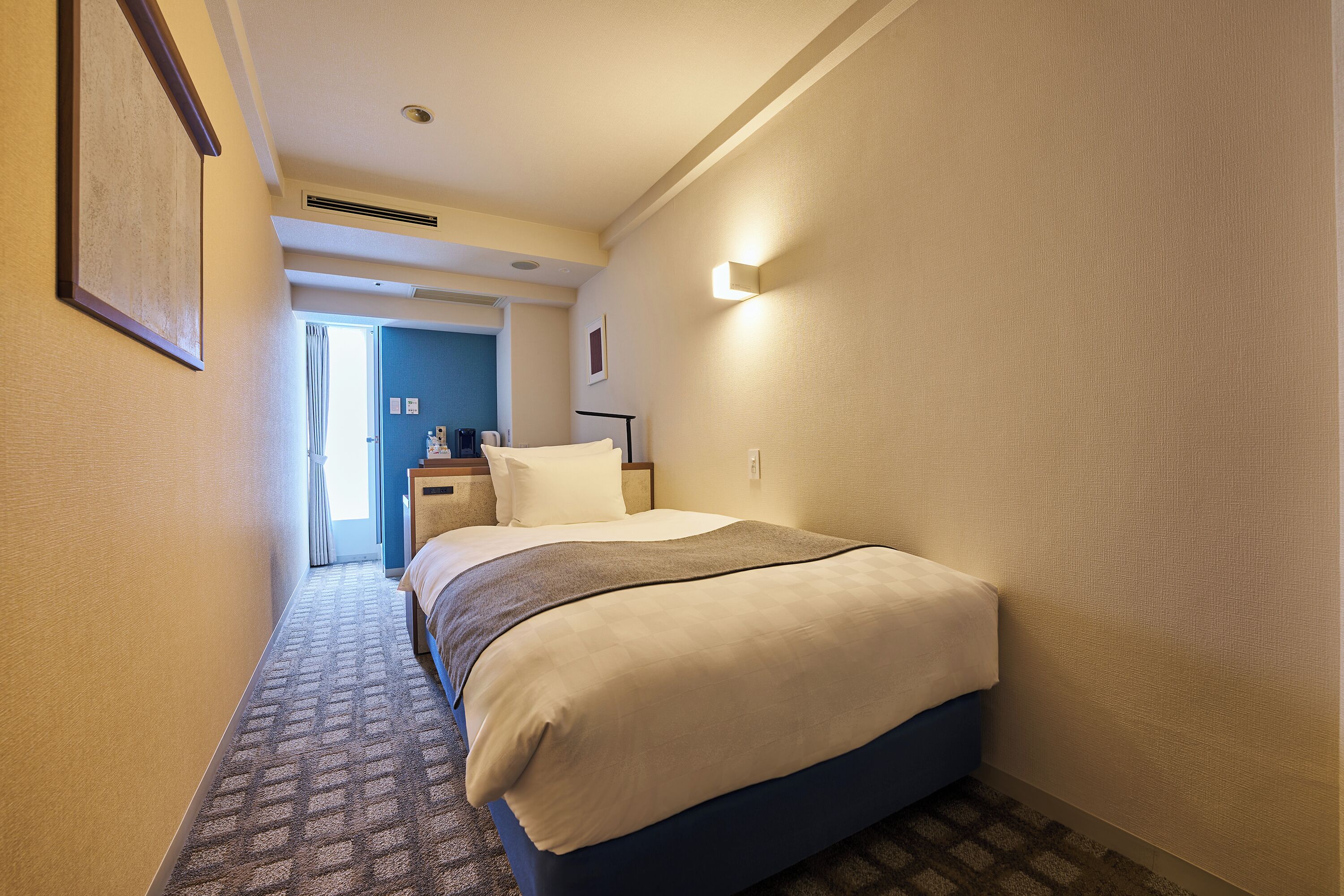 A hotel room with a bed, a nightstand, a wall-mounted light, and a door leading to another room.