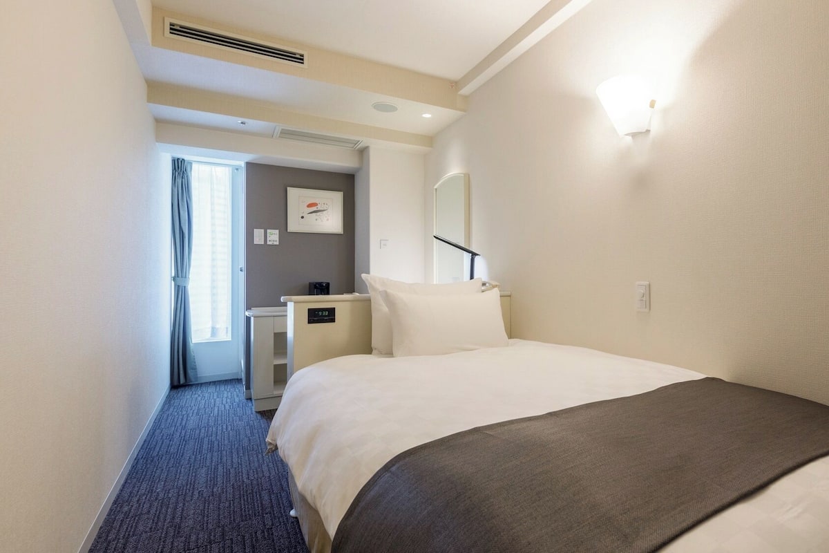 A hotel room with a bed, a desk, a chair, a mirror, and a door leading to another room.