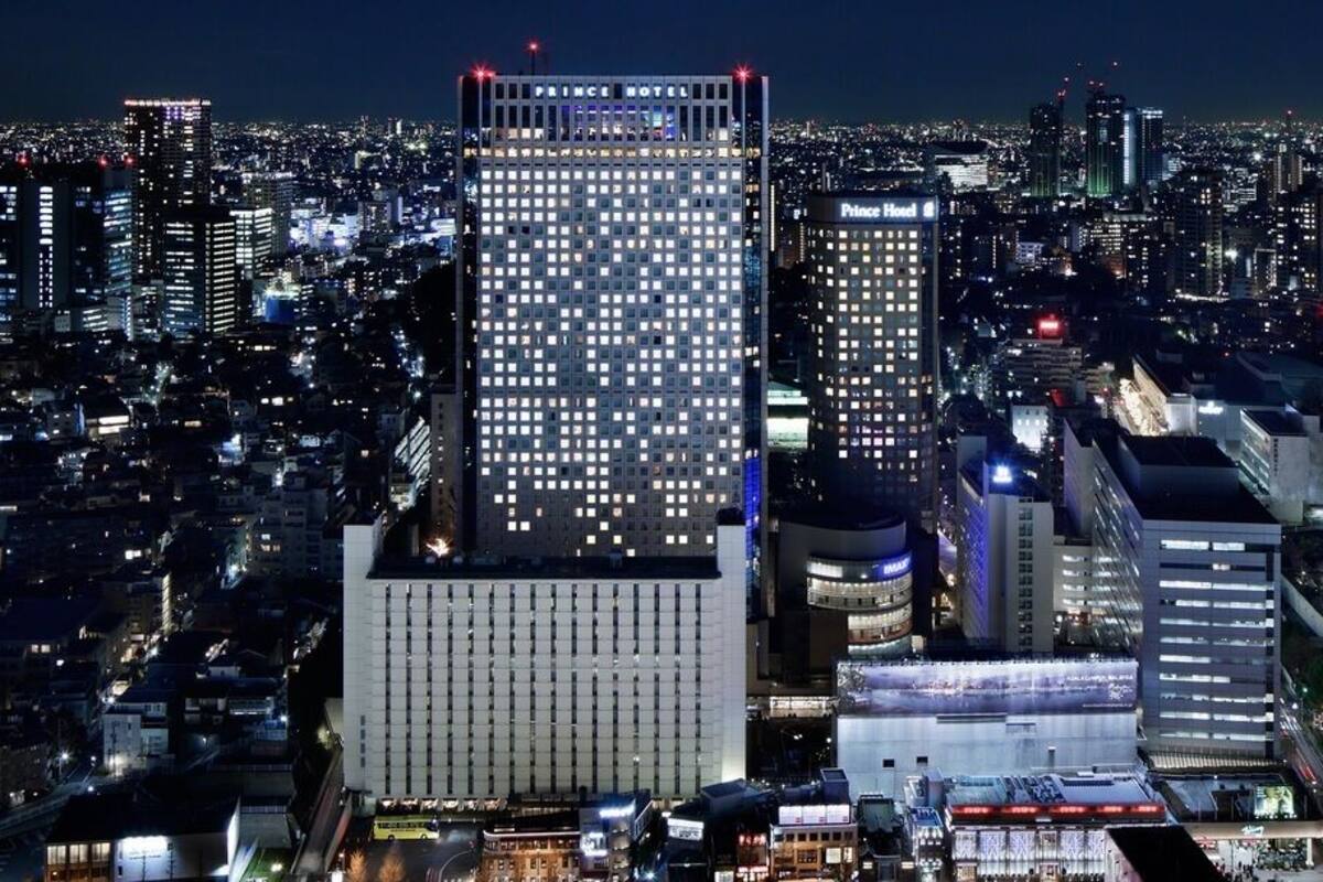 Une vue nocturne d’une ville, avec des bâtiments imposants, notamment l’hôtel Prince et un grand hôtel dont la façade est constituée d’une grille de fenêtres.