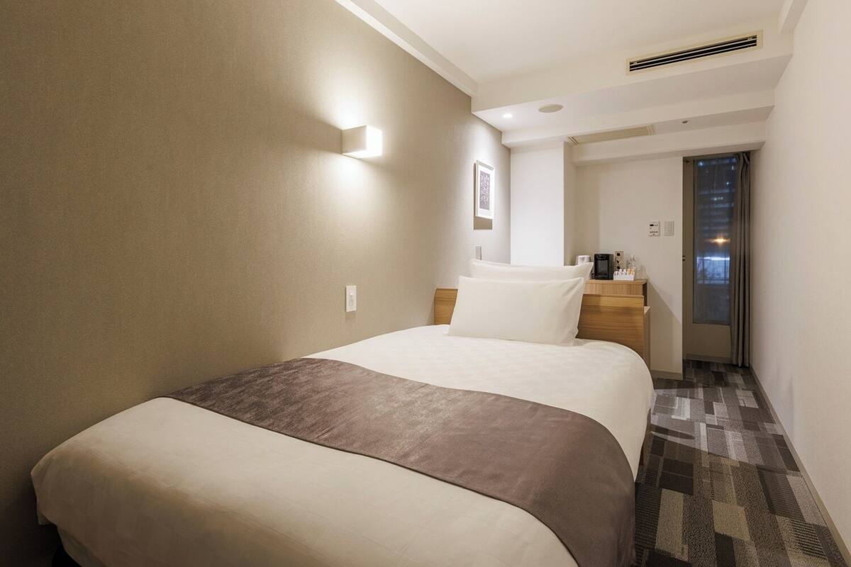 Une chambre d’hôtel comprenant un lit, une table de chevet, une lampe fixée au mur et une porte donnant sur une autre pièce.