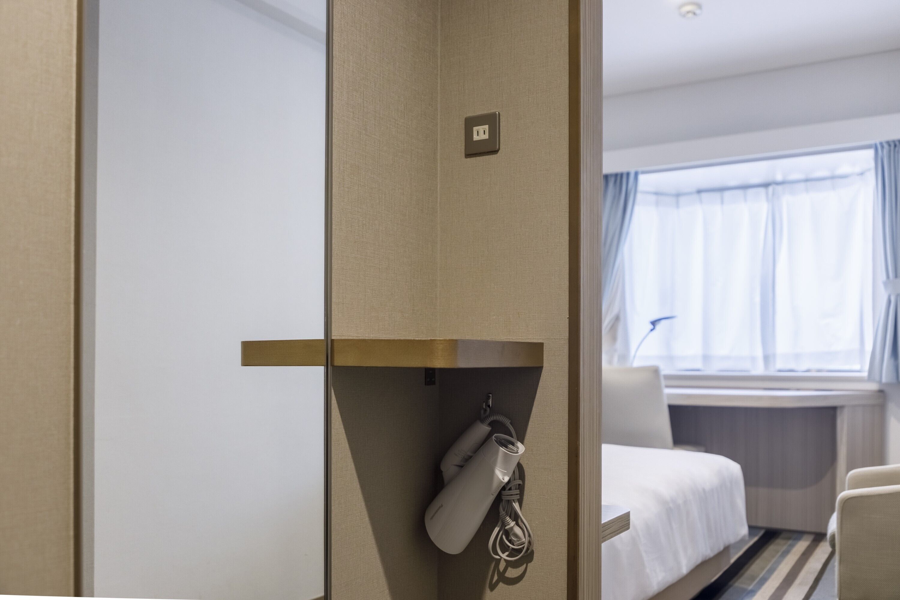 A hotel room with a bed, a desk, a chair, a window with curtains, and a wall-mounted phone.