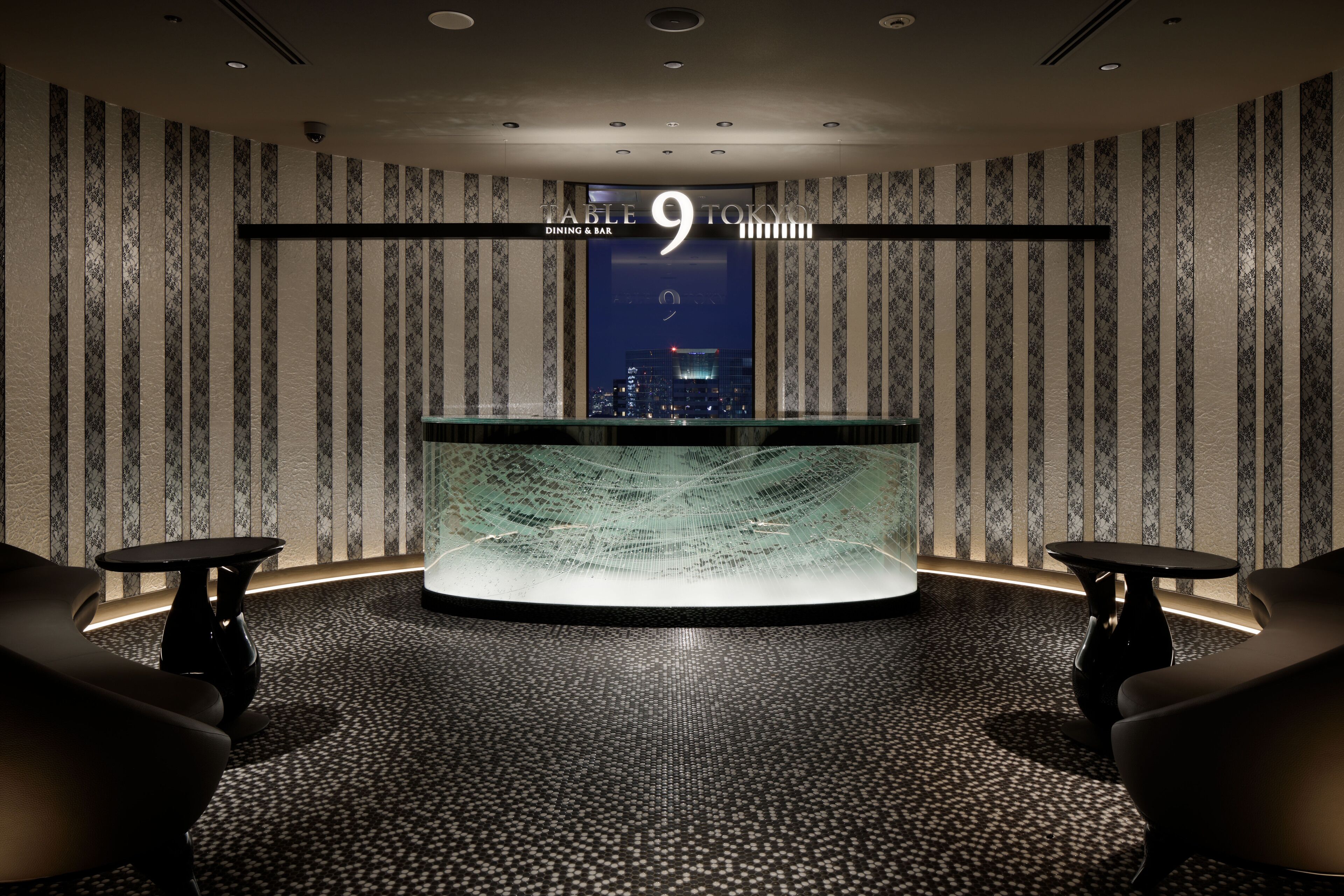 Un intérieur moderne avec une table centrale et deux chaises, une vue sur le paysage urbain par une fenêtre, et une enseigne indiquant « Table 9 Tokyo Dining & Bar ».