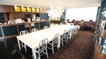 A spacious dining area with a long white table and chairs, a bar area with a coffee machine, and a seating area with a brown sofa and patterned carpet.