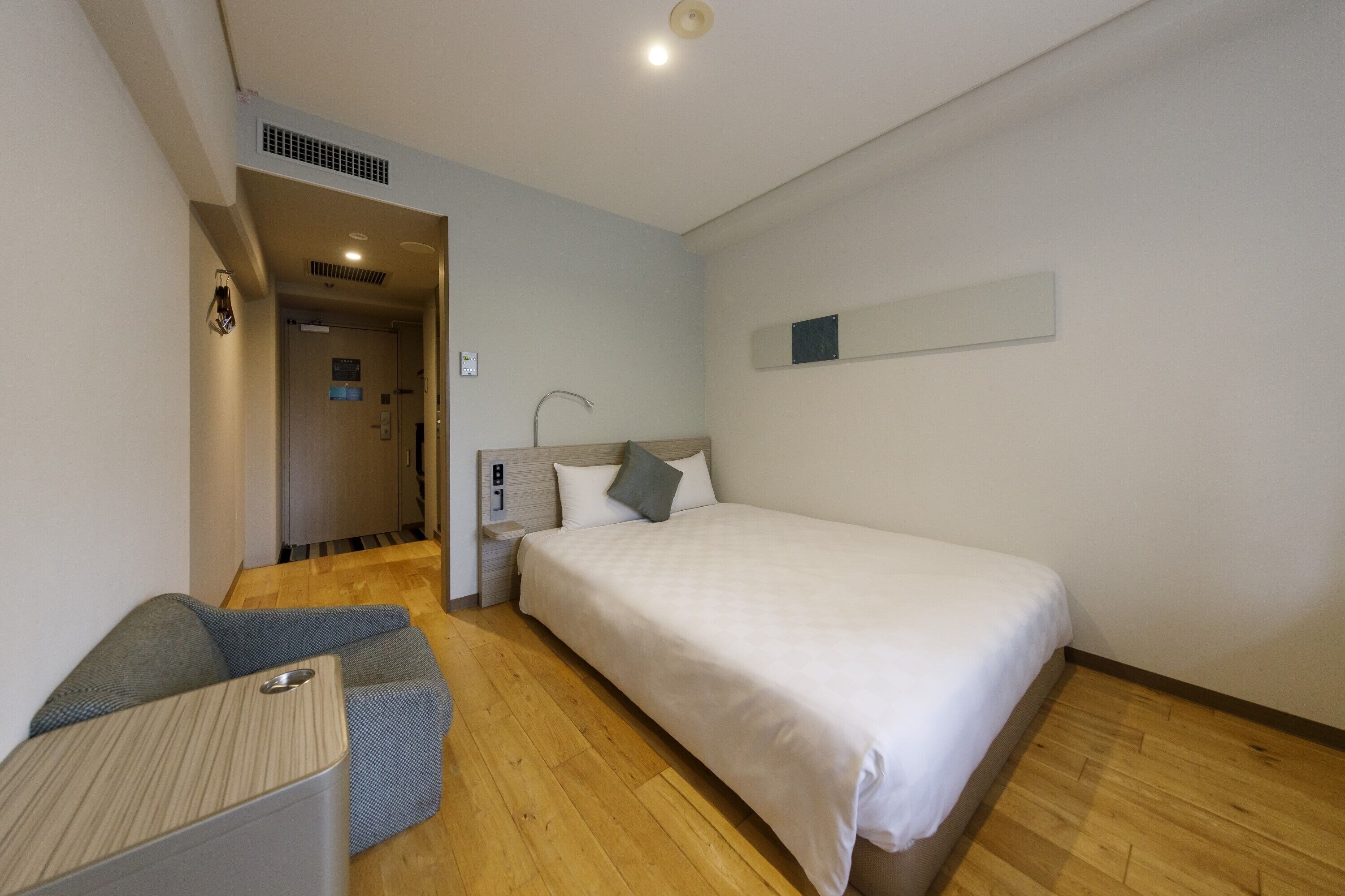 A hotel room with a bed, a small desk, and a chair. There is a door leading to another room and a vent on the wall.
