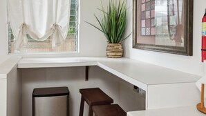 A corner of a room with a white countertop, a trash bin, a wooden stool, a potted plant, a window with sheer curtains, and a framed picture on the wall.