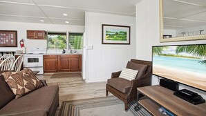 A living room with a brown sofa, a television, and a dining table. There is a kitchen area with wooden cabinets and a white refrigerator.