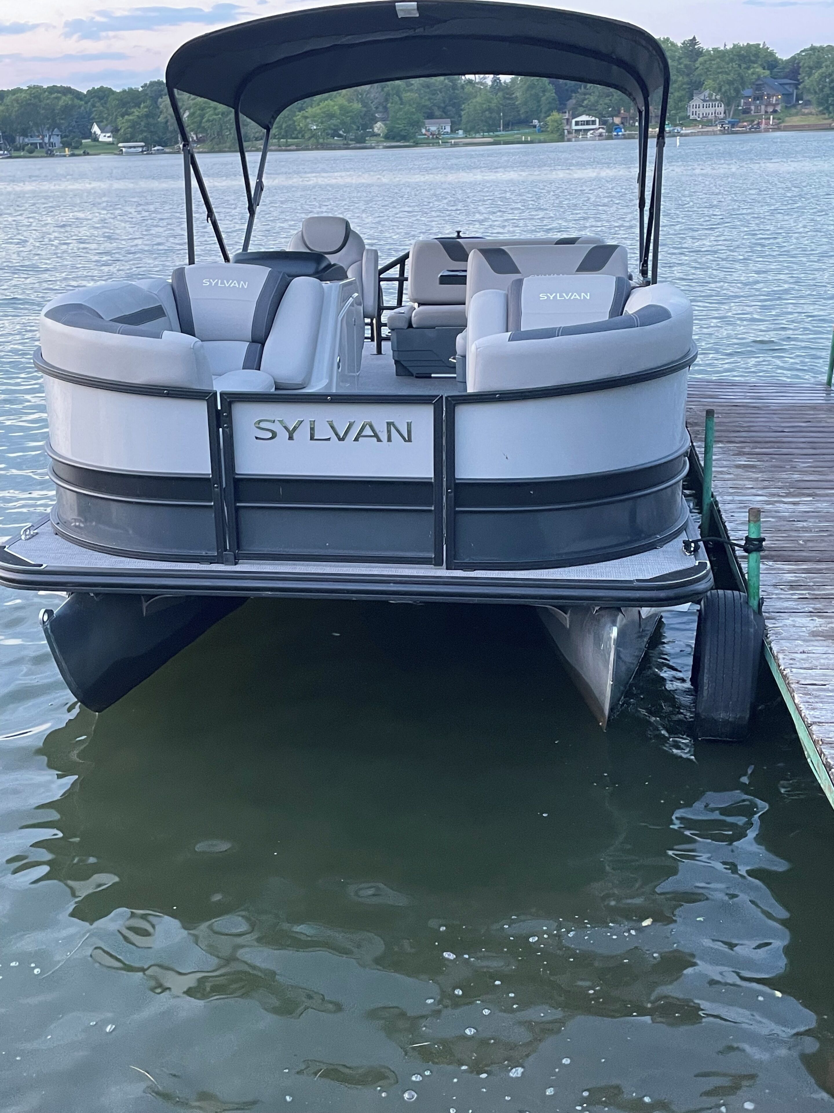 A boat with a brand name 'SYLVAN' on the back, docked at a pier with calm water and houses in the background.