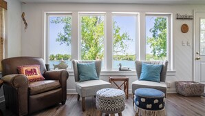 Un coin salon accueillant avec un fauteuil en cuir marron et deux fauteuils, chacun agrémenté d’un coussin bleu. On y trouve également deux poufs avec des coussins à motifs. De grandes fenêtres offrent une vue sur les arbres et un plan d’eau.
