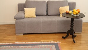 A grey sofa with two yellow cushions, a small round table with a fruit bowl, and a patterned rug on a wooden floor. - 270 Pulawska (Warsaw)