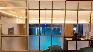 Une pièce dotée d’une grande fenêtre donnant sur une piscine, d’un plancher en bois et d’une petite table ornée d’une plante et de bouteilles.