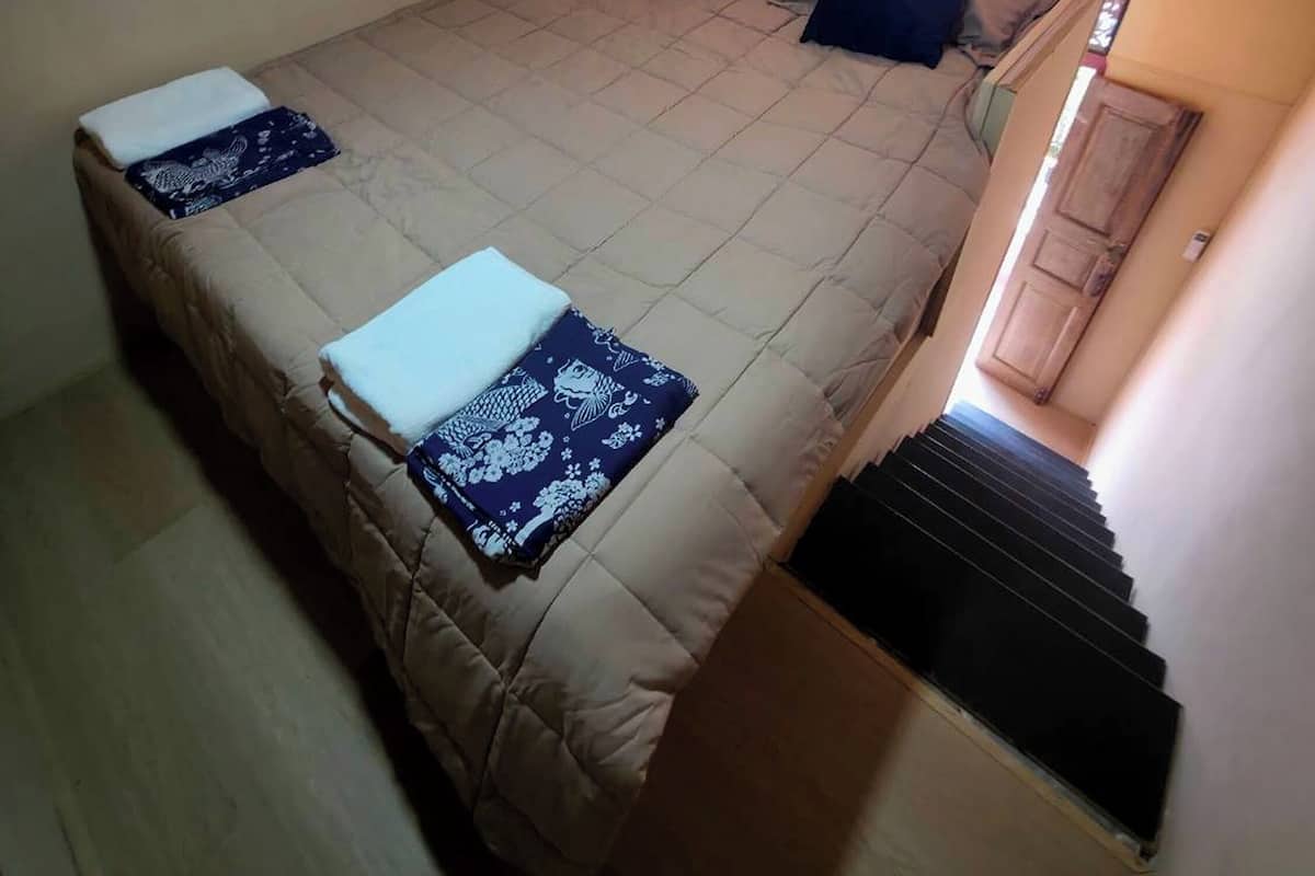 A single-bed room with a beige bedspread, two pillows, and a folded blanket. There is a window with blinds, a small table with a folded blanket, and a staircase leading to an upper level.