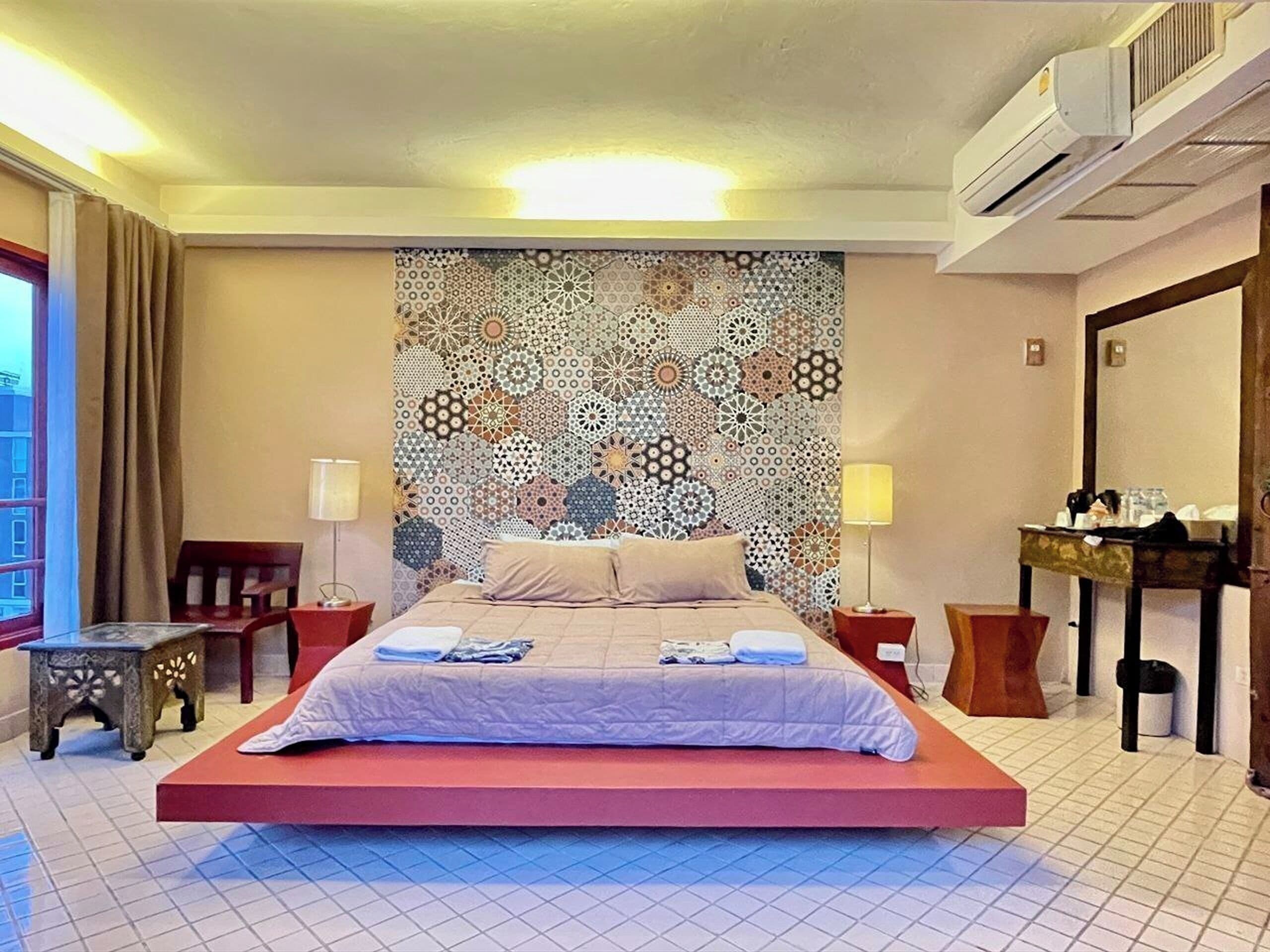 Quarto de hotel com cama, cabeceira estampada, mesa de cabeceira, cadeira, pequena mesa e unidade de ar condicionado.