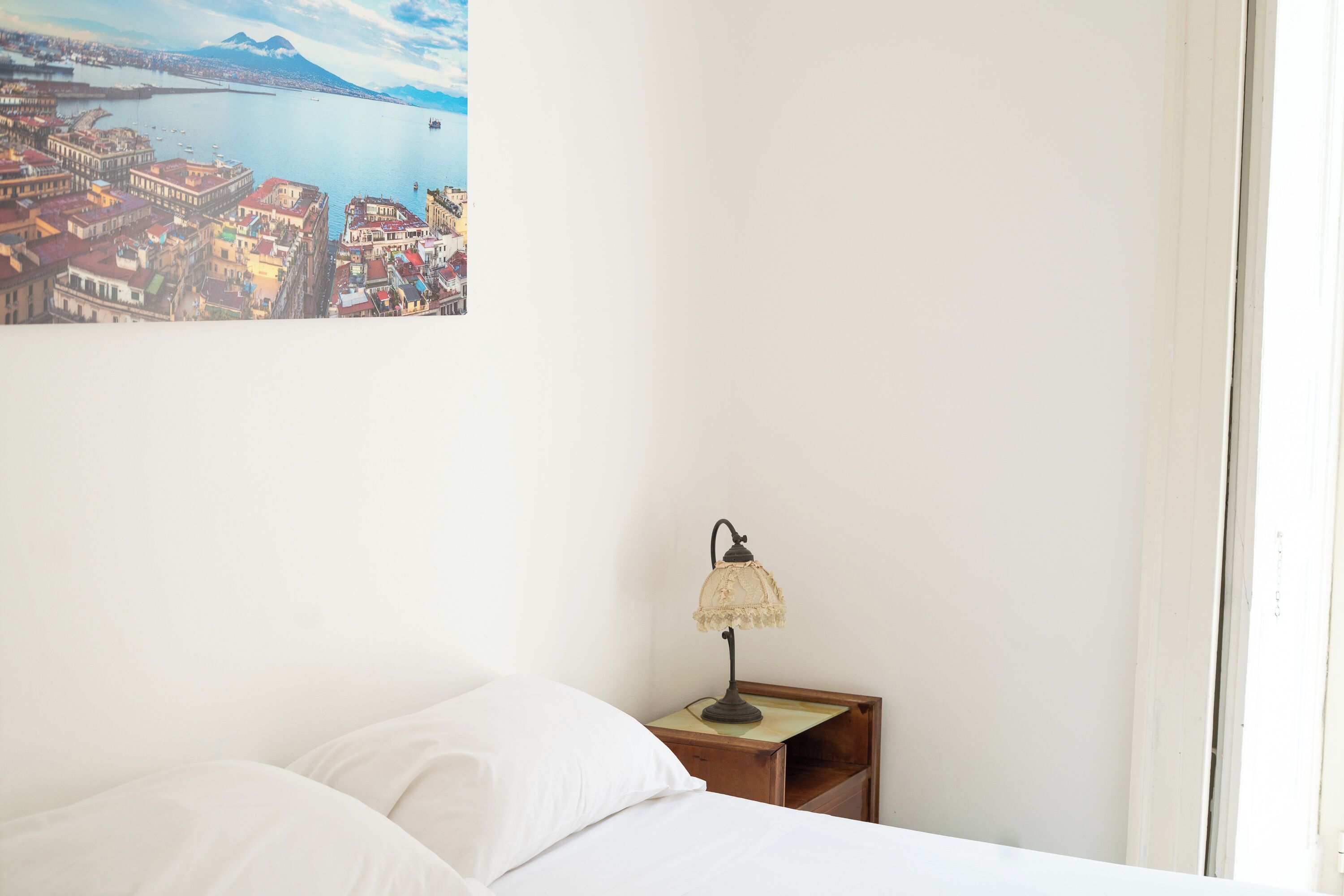 A bedroom with a bed, a nightstand, a lamp, and a painting of a coastal cityscape.