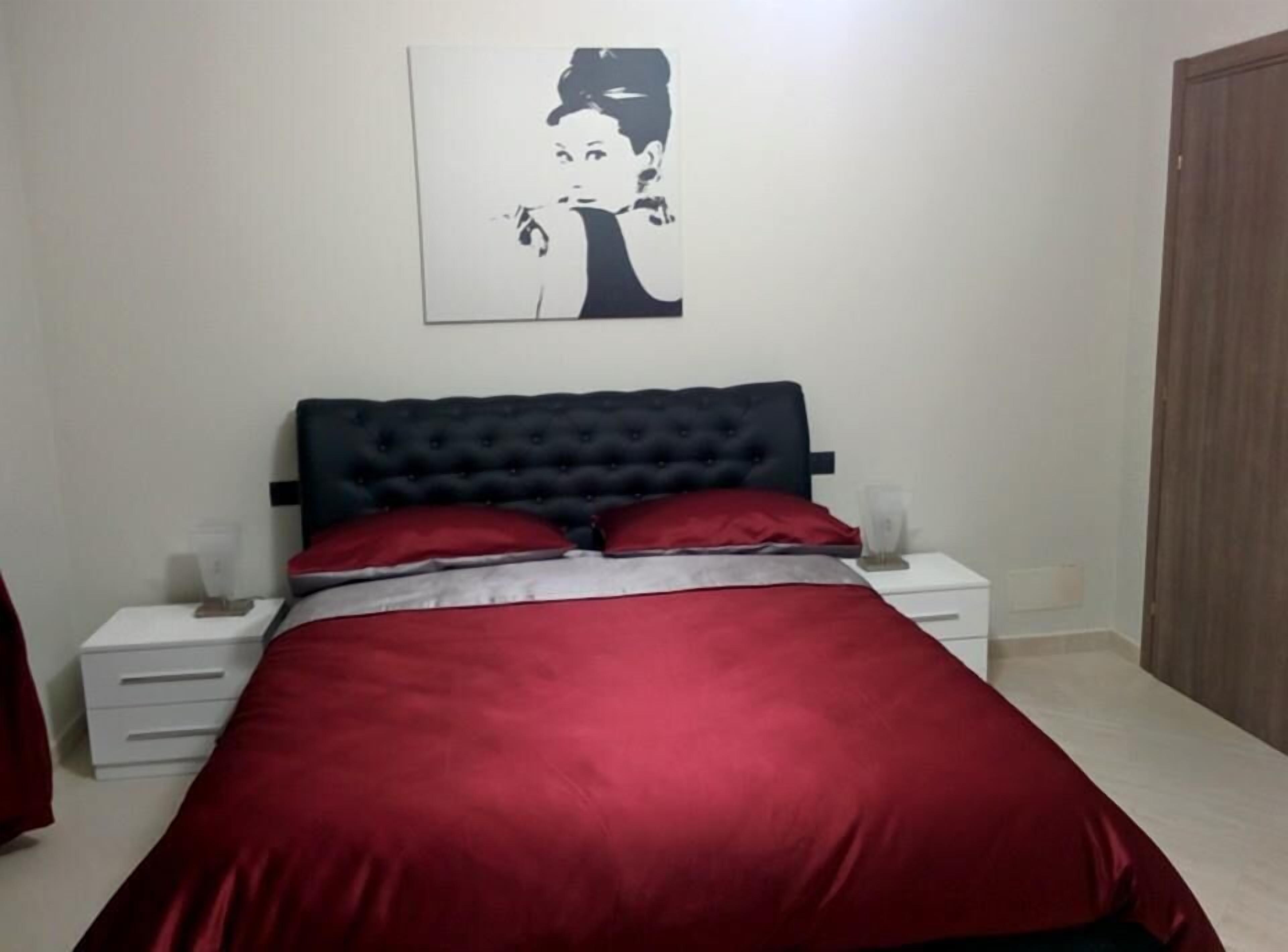 A bedroom with a dark headboard, red bedding, a white bedside table, and a framed picture on the wall.