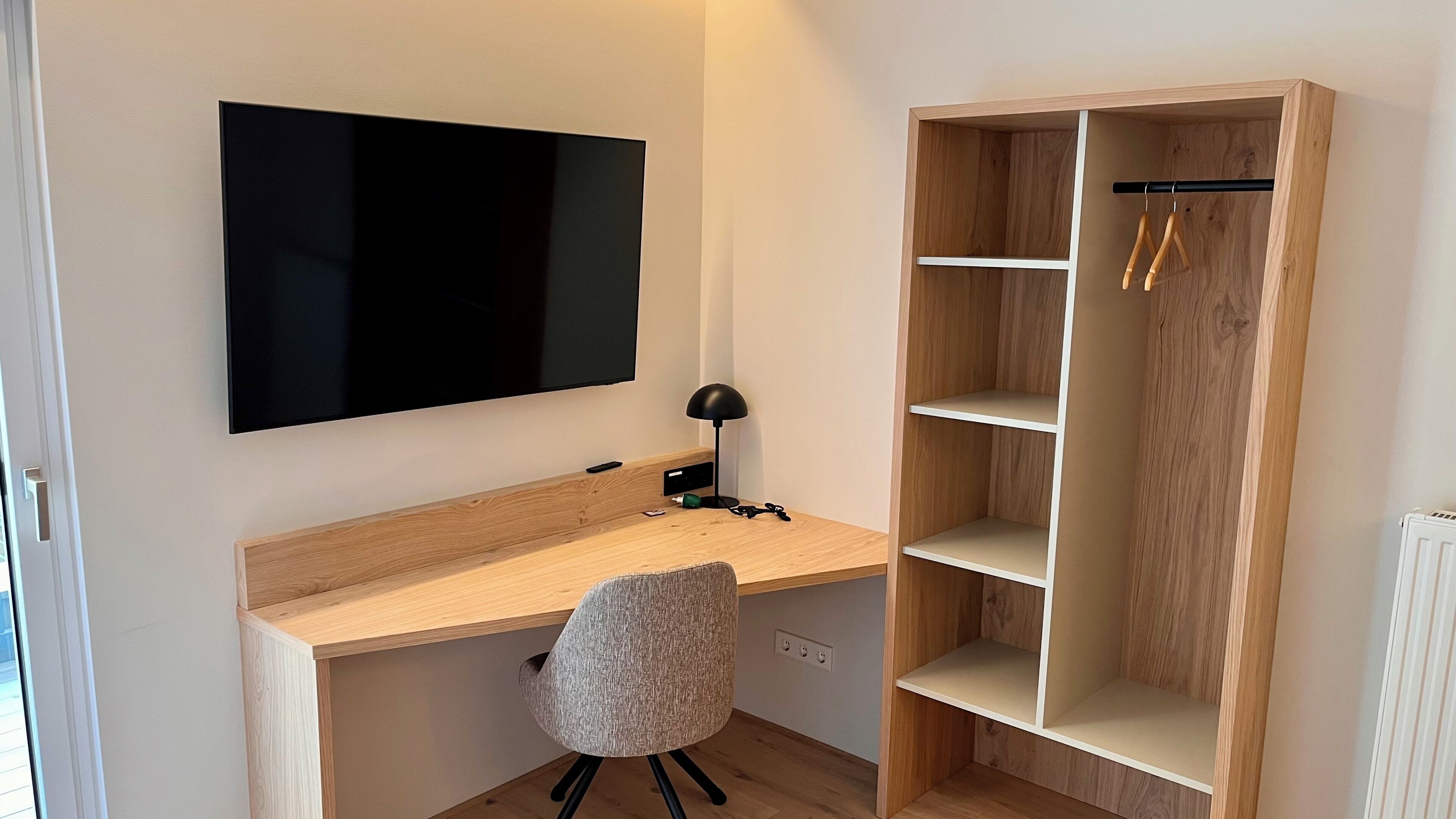 Una habitación con un escritorio de madera, una silla, un televisor montado en la pared y una estantería de madera.