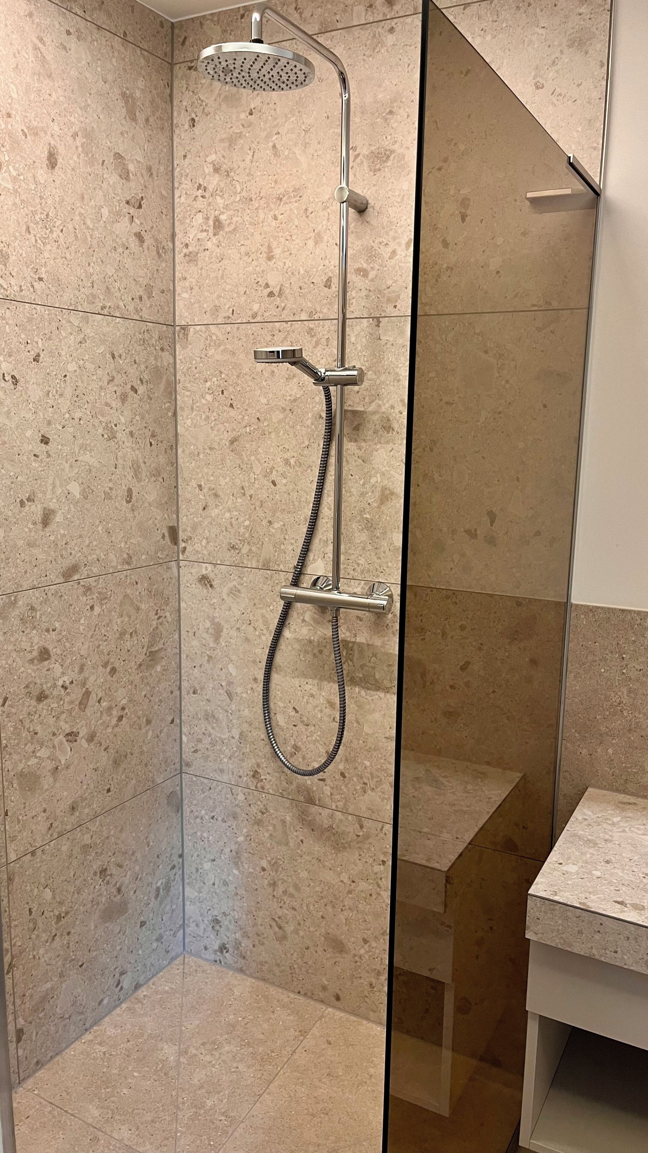 A shower with a handheld showerhead and a rain shower head, a glass door, and a tiled wall.