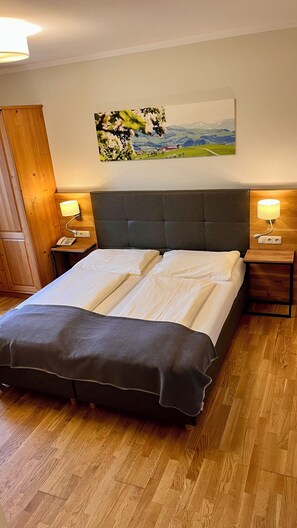 A bed with a grey headboard, two bedside tables with lamps, a wooden floor, and a painting of a landscape on the wall. - Gafringwirt (Euratsfeld)