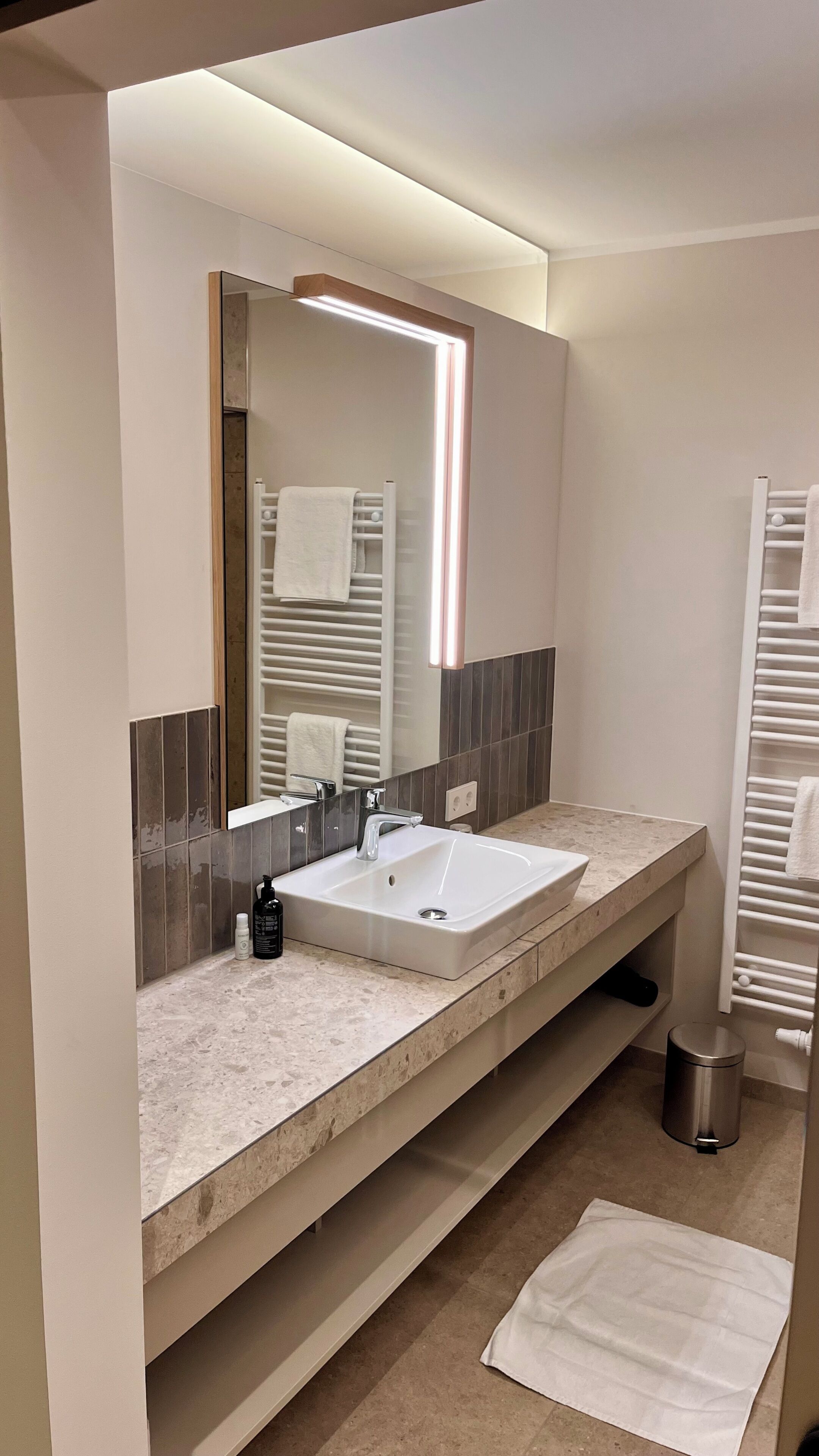 Un baño moderno con un espejo grande, dos lavamanos, un perchero para toallas y un cubo de basura.