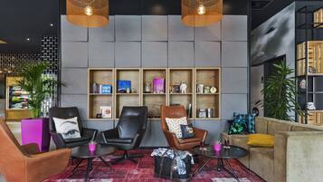 A modern lounge area with leather chairs, a sofa, and a red patterned rug. There are shelves with books and decorative items, and two hanging lamps.