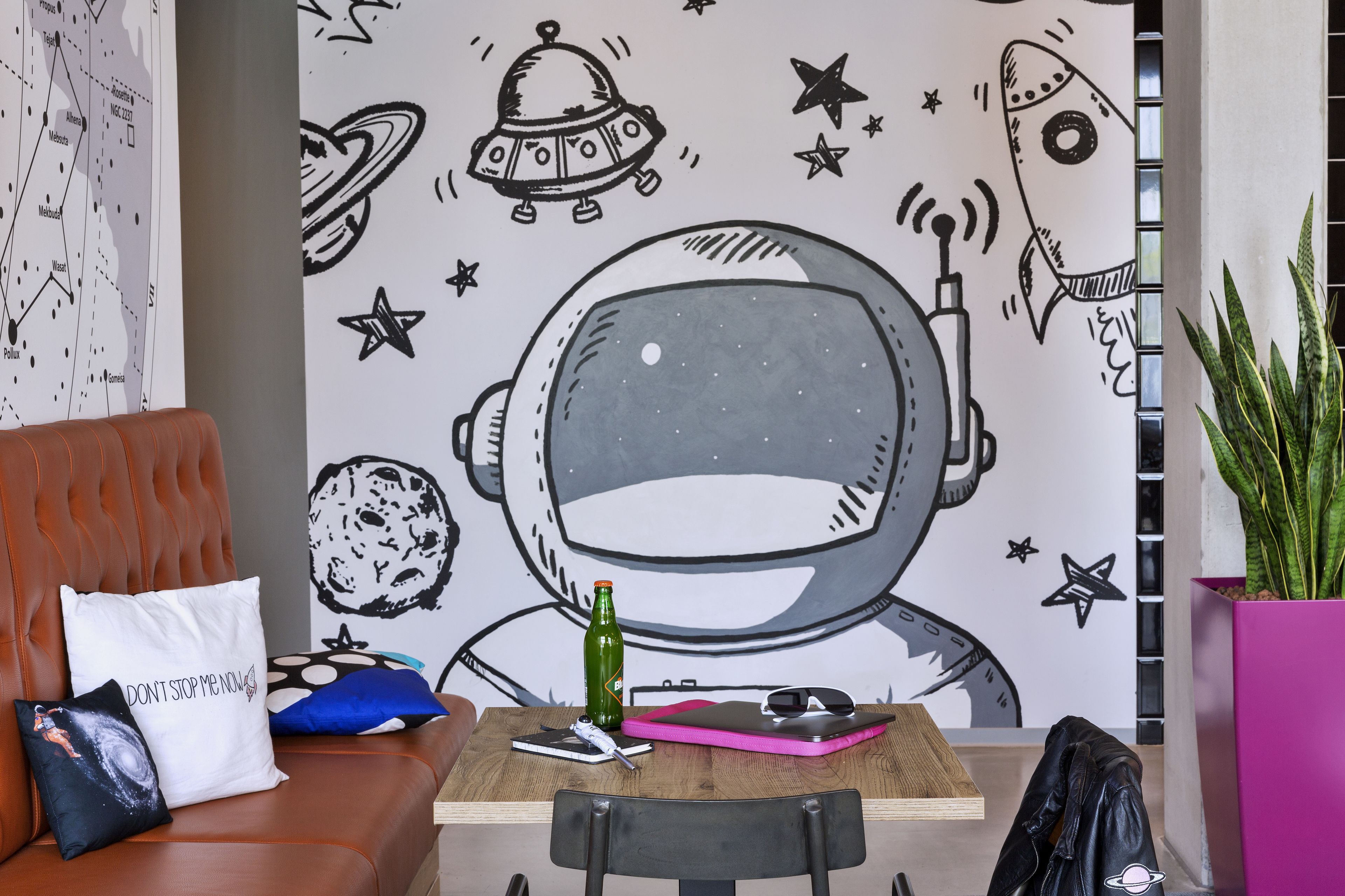 A room with a large wall mural of an astronaut, a brown leather couch, a wooden table with a laptop, a green bottle, and a pink notebook.