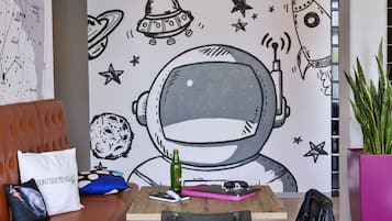 A room with a large wall mural of an astronaut, a brown leather couch, a wooden table with a laptop, a green bottle, and a pink notebook.