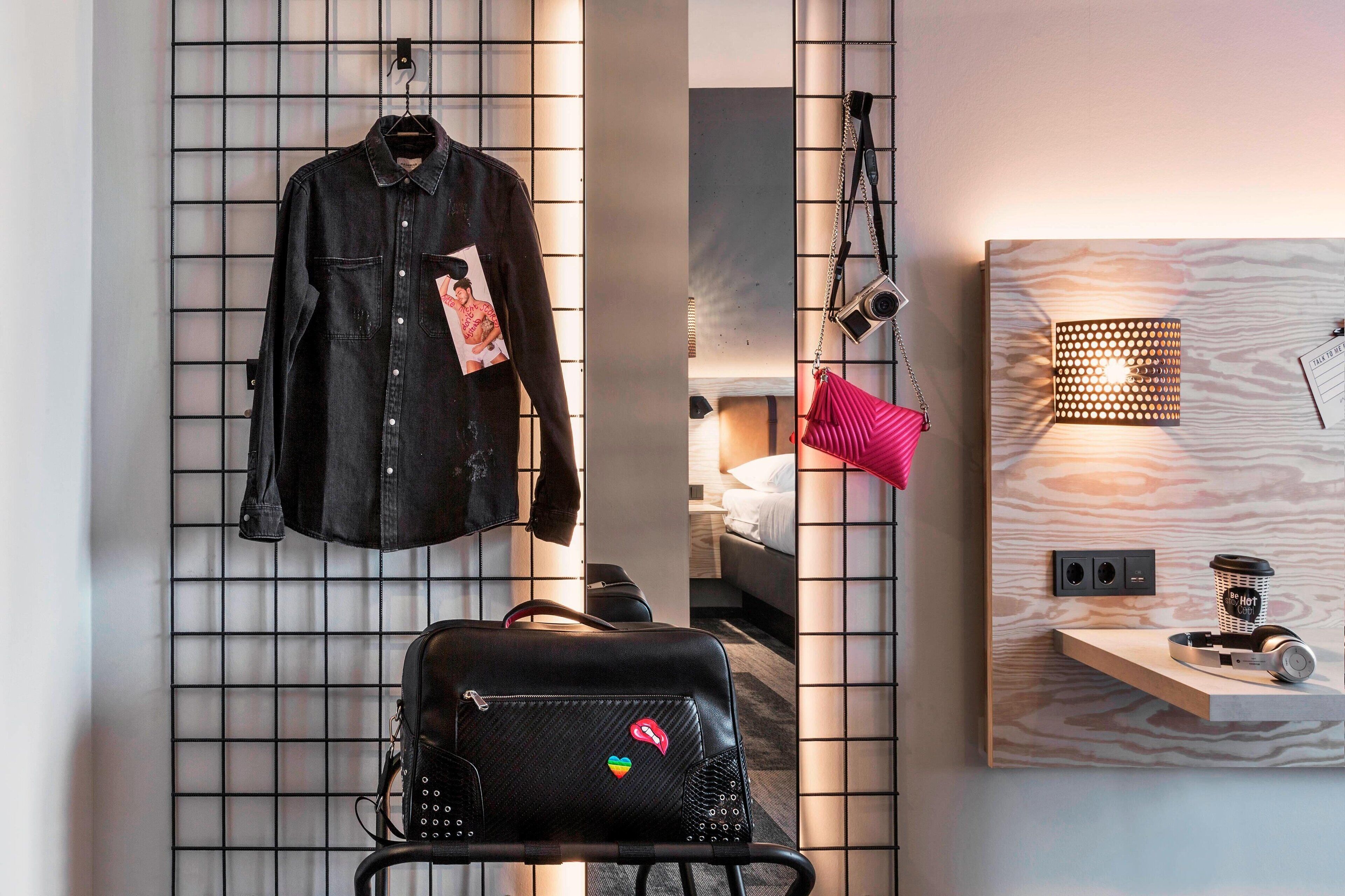 Habitación de hotel con una cama, un bolso colgado, una chaqueta y una estantería con lámpara y cámara.