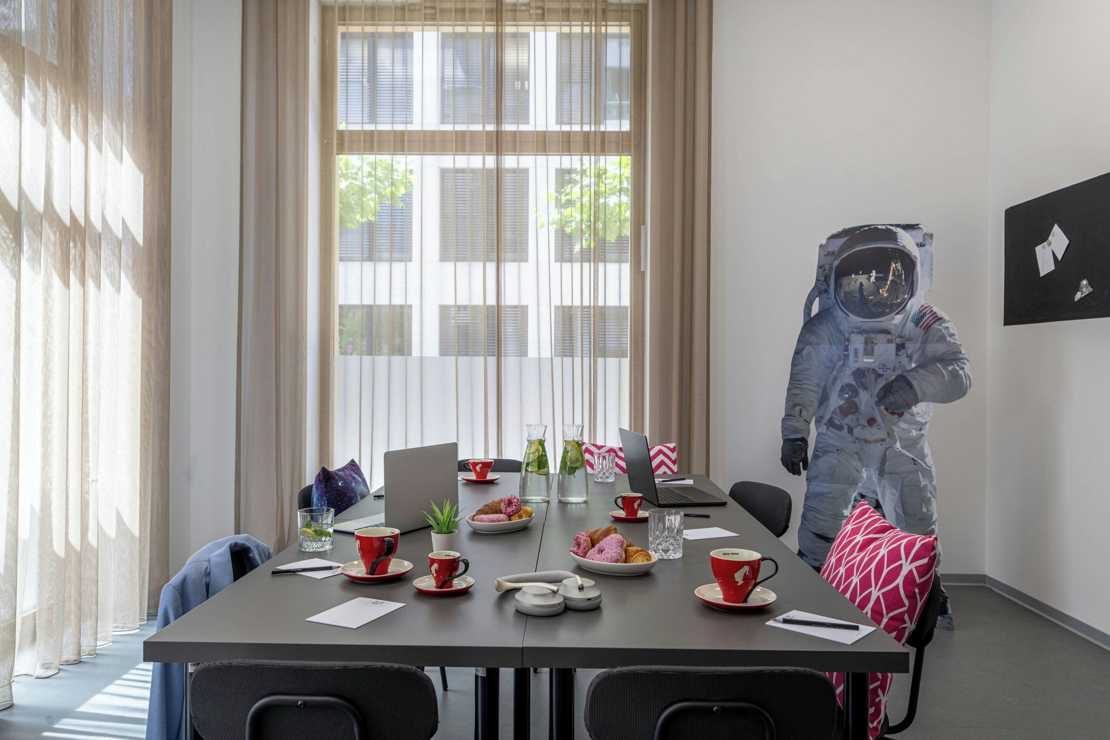Une salle de conférence avec une table préparée pour une réunion, un ordinateur portable et une personne vêtue d’un costume d’astronaute.