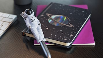 A notebook with a space-themed cover, a pen, and a small astronaut figurine on a wooden surface.