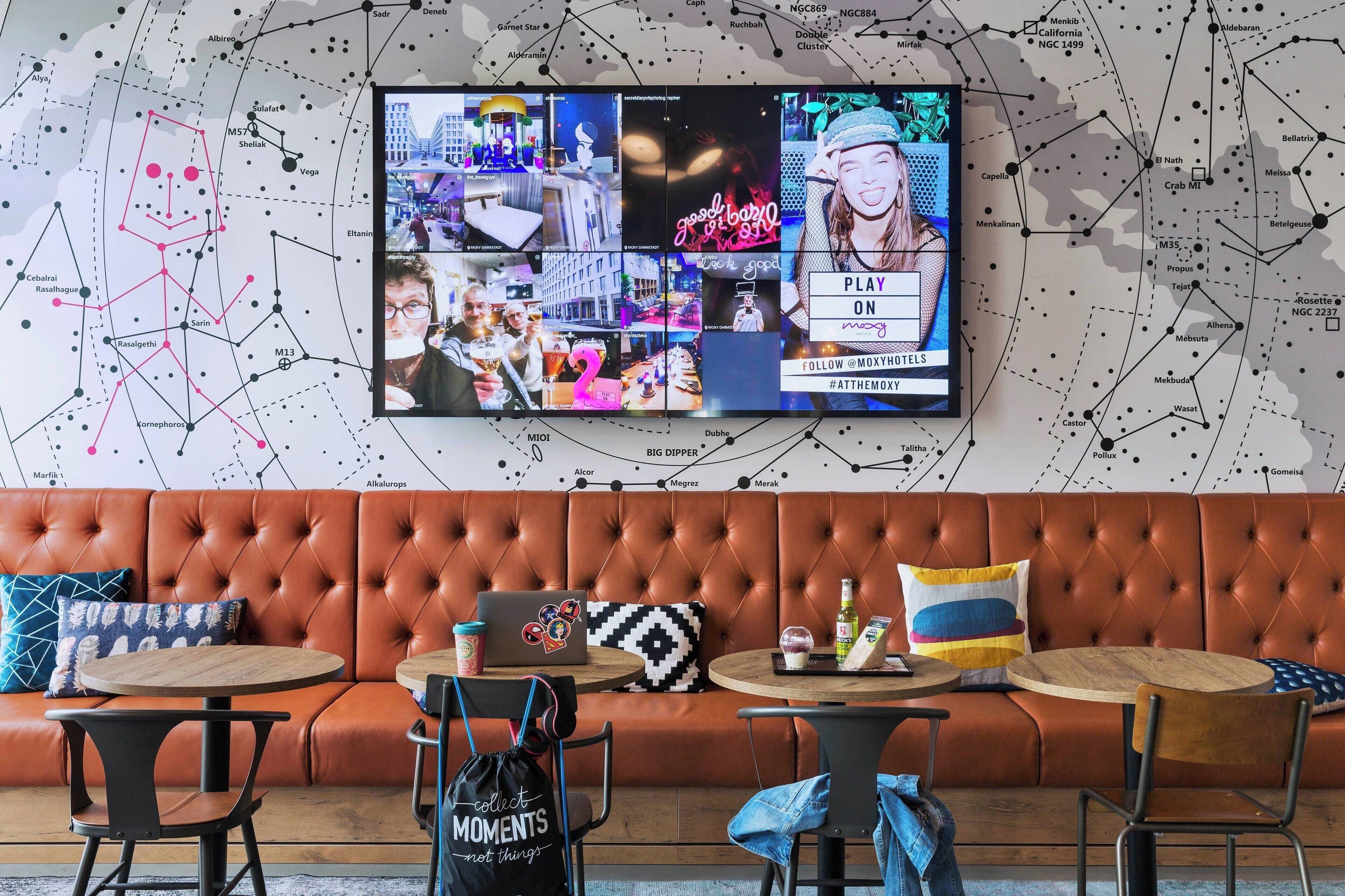 Zona d'estar moderna amb un televisor gran de pantalla penjada que mostra diverses imatges, un sofà de pell, taules rodones i una màquina de cafè.