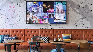 Zona d'estar moderna amb un televisor gran de pantalla penjada que mostra diverses imatges, un sofà de pell, taules rodones i una màquina de cafè.