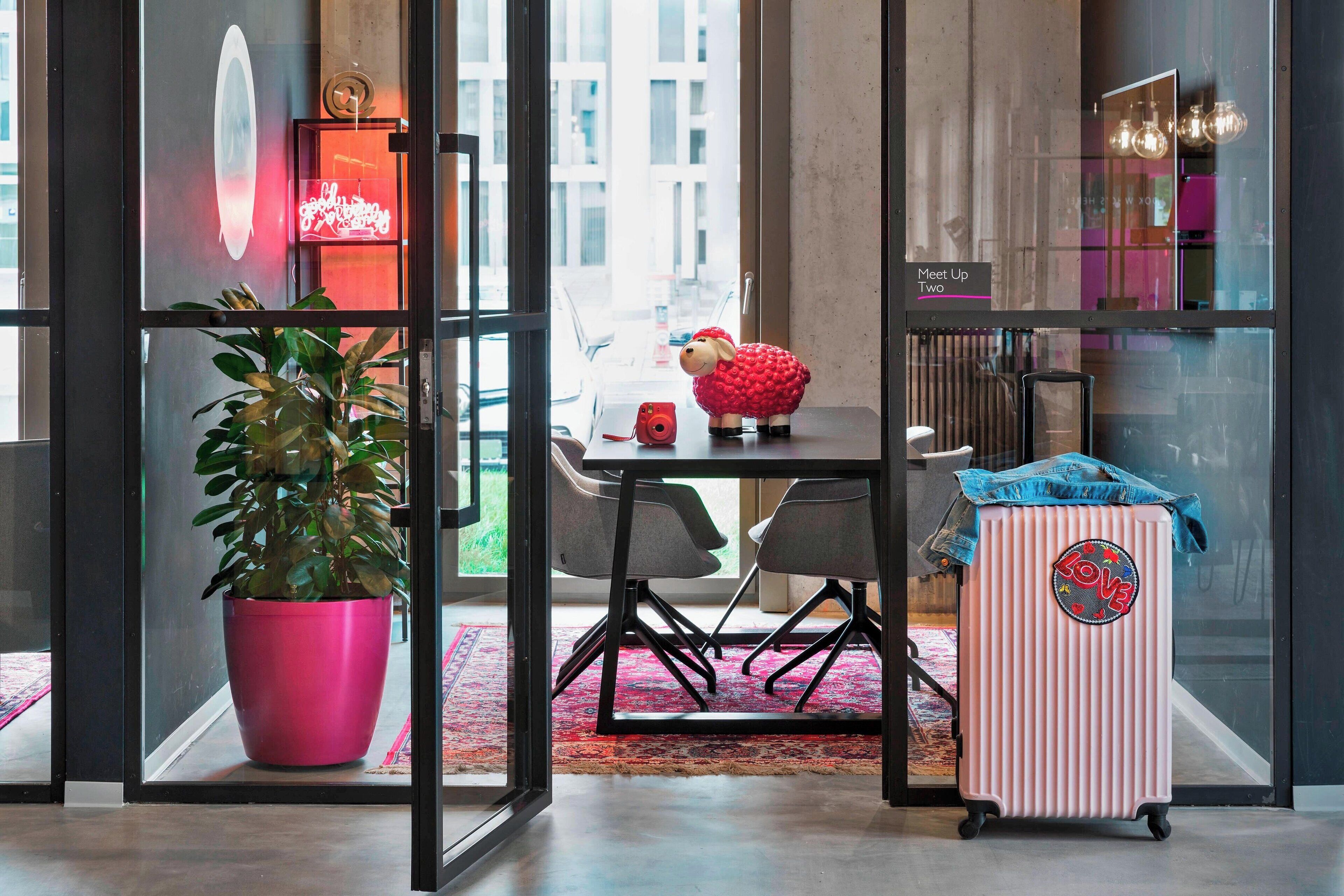 A modern interior with a glass partition, a potted plant, a table with chairs, a suitcase, and a neon sign.