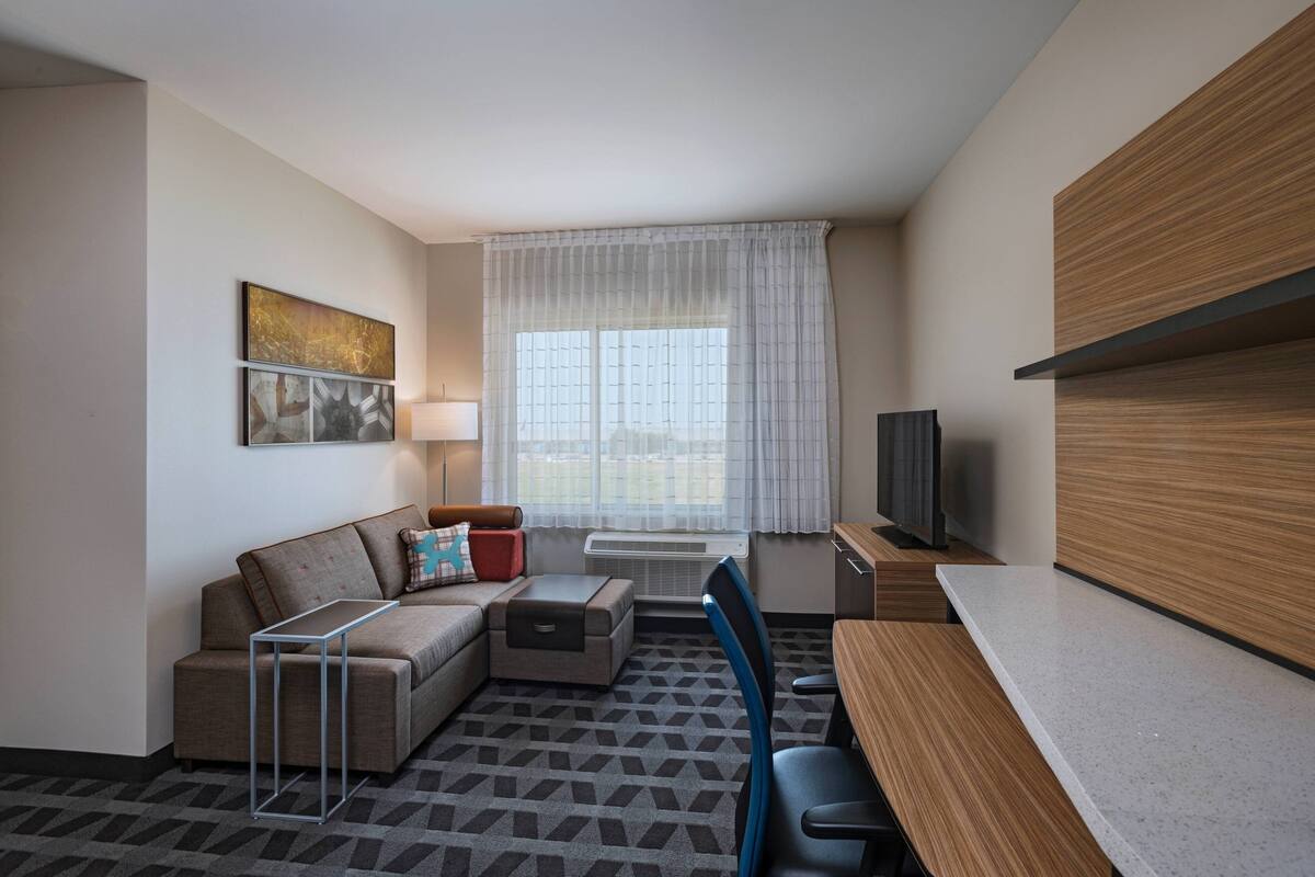 Ein modernes Hotelzimmer mit einer Couch, einem kleinen Tisch, einem Fernseher und einem Fenster mit Vorhängen.