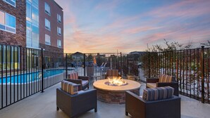 An outdoor patio with wicker furniture and a central fire pit, surrounded by a pool and a fence.