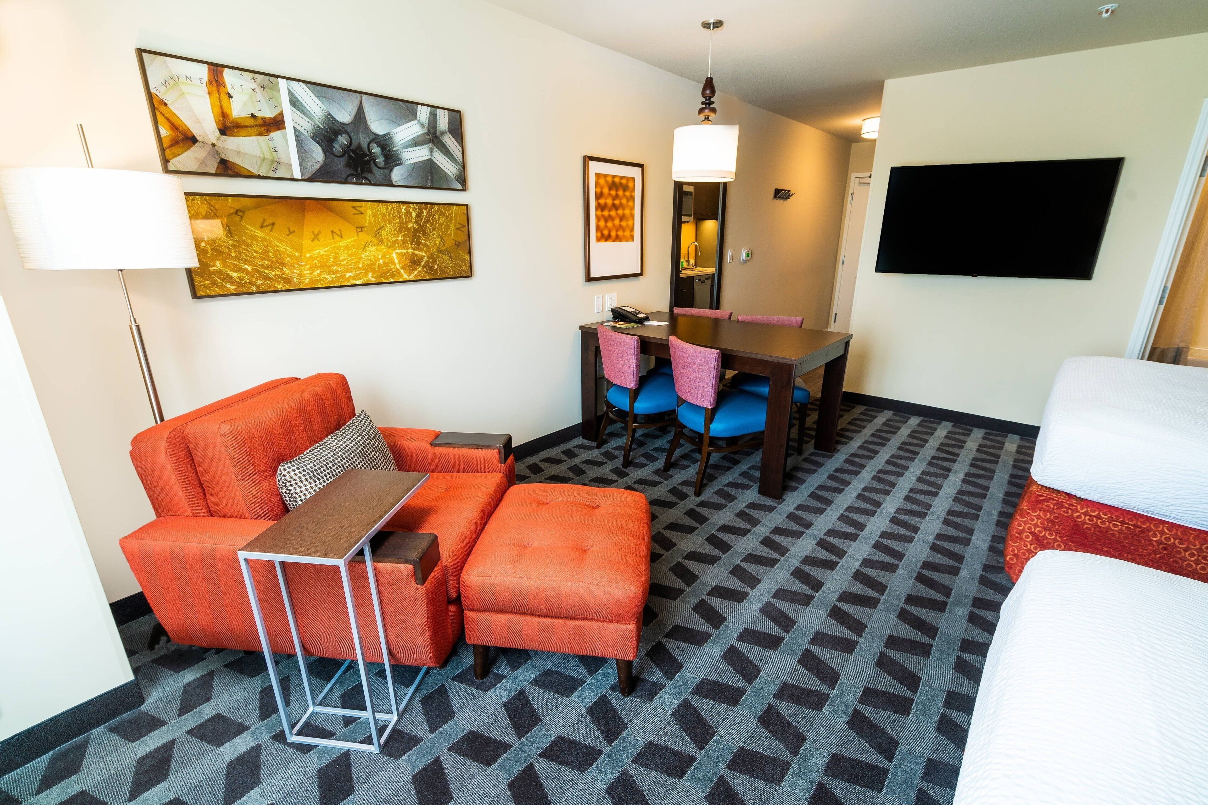 A hotel room with a sofa, ottoman, table, chairs, a TV, and artwork on the walls.