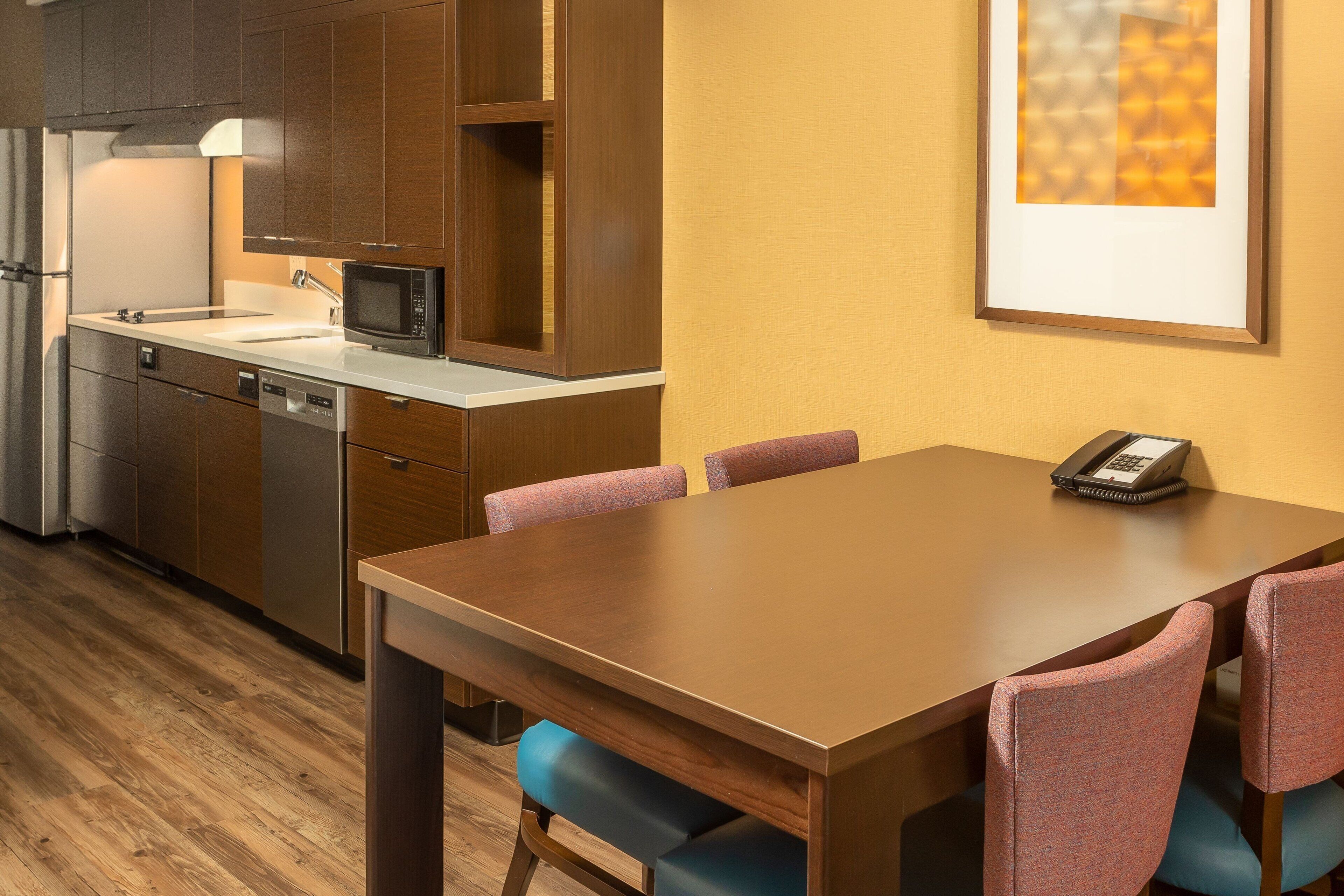 A modern kitchen with wooden cabinets, a microwave, and a sink. There is a dining table with chairs and a phone on it.