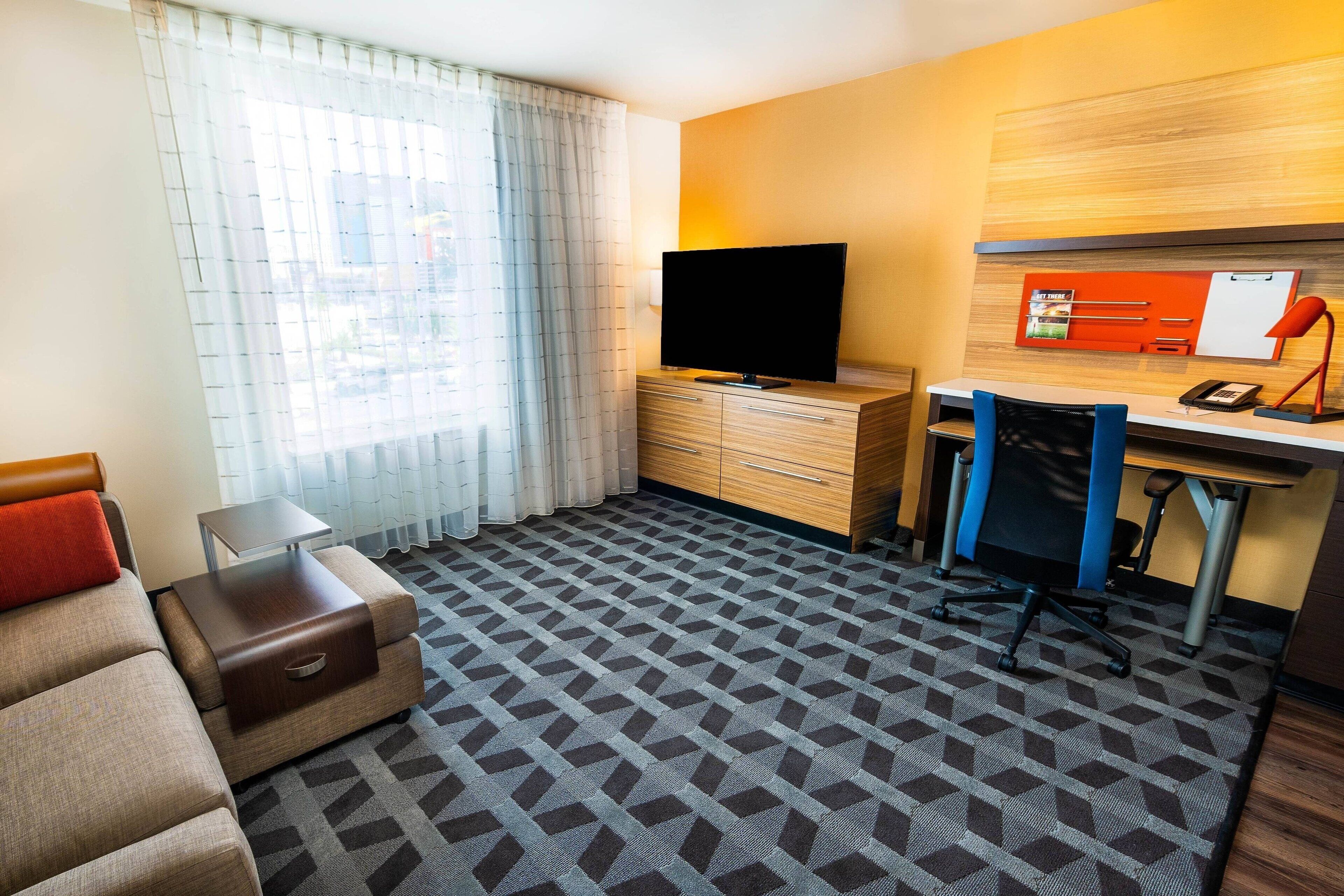 A hotel room with a sofa, a desk with a computer, a television, and a patterned carpet.