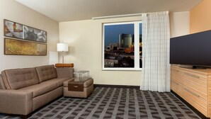 A hotel room with a sofa, a small table, a lamp, a TV, and a window with a city view. - TownePlace Suites By Marriott Las Vegas Stadium District (Las Vegas)