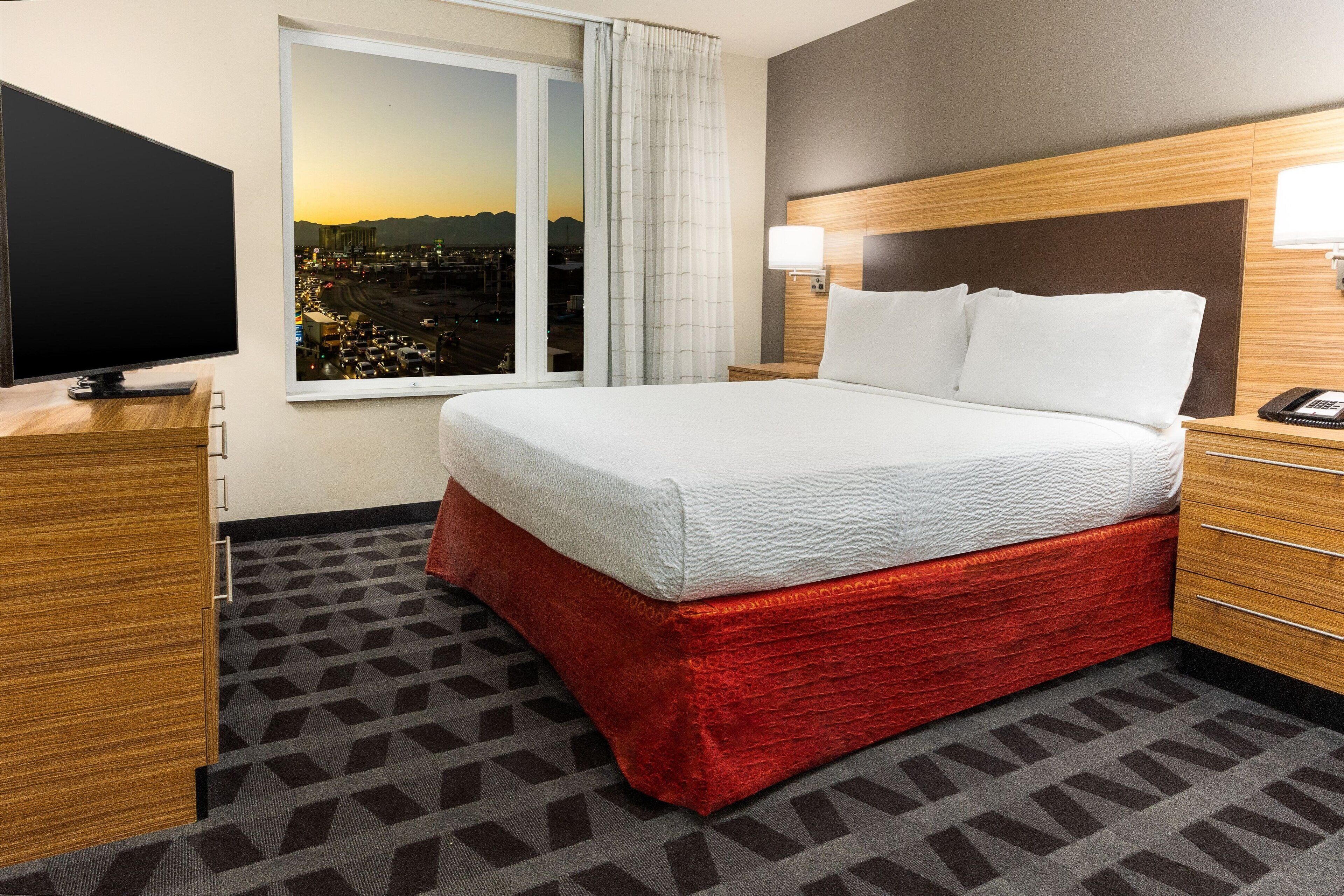 A hotel room with a large bed, a TV, a window with a city view, and a wooden headboard.